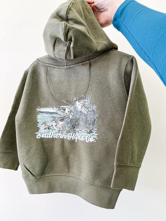 A hand holds up an olive green Pintails Skies Hoodie by Southern Hyde Co, featuring an outdoor scene with ducks and reeds on the back.
