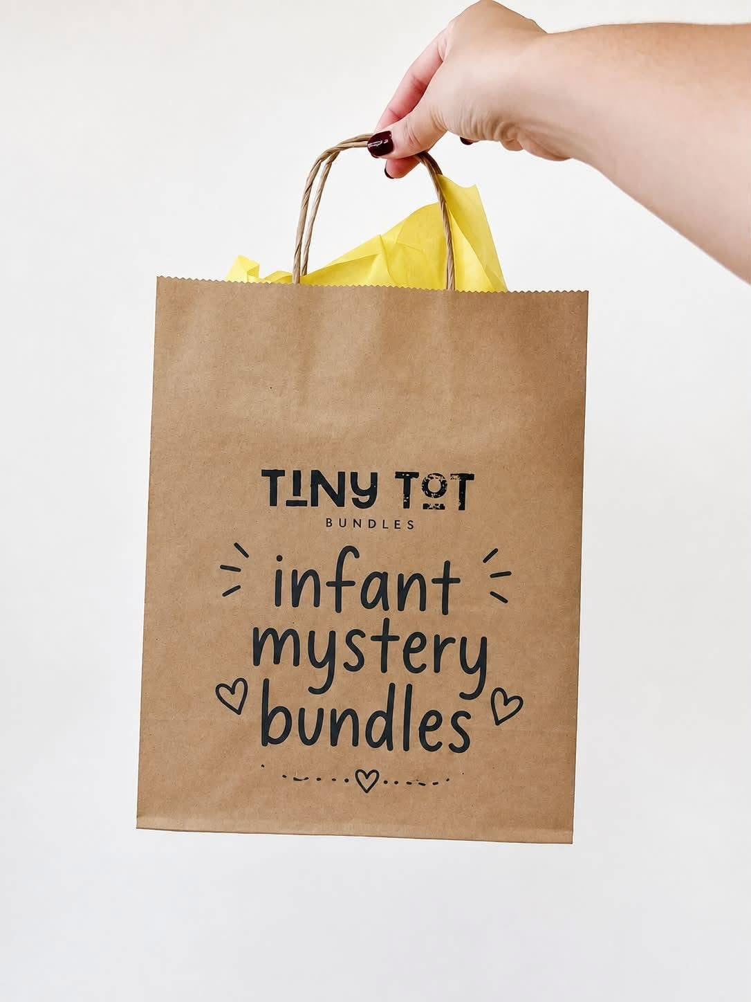 A hand holds a brown paper gift bag labeled with the Tiny Tot Bundles $15 Mystery Bundle. Yellow tissue paper peeks out of the bag, emphasizing that each bundle is a final sale and includes items all in the same size.