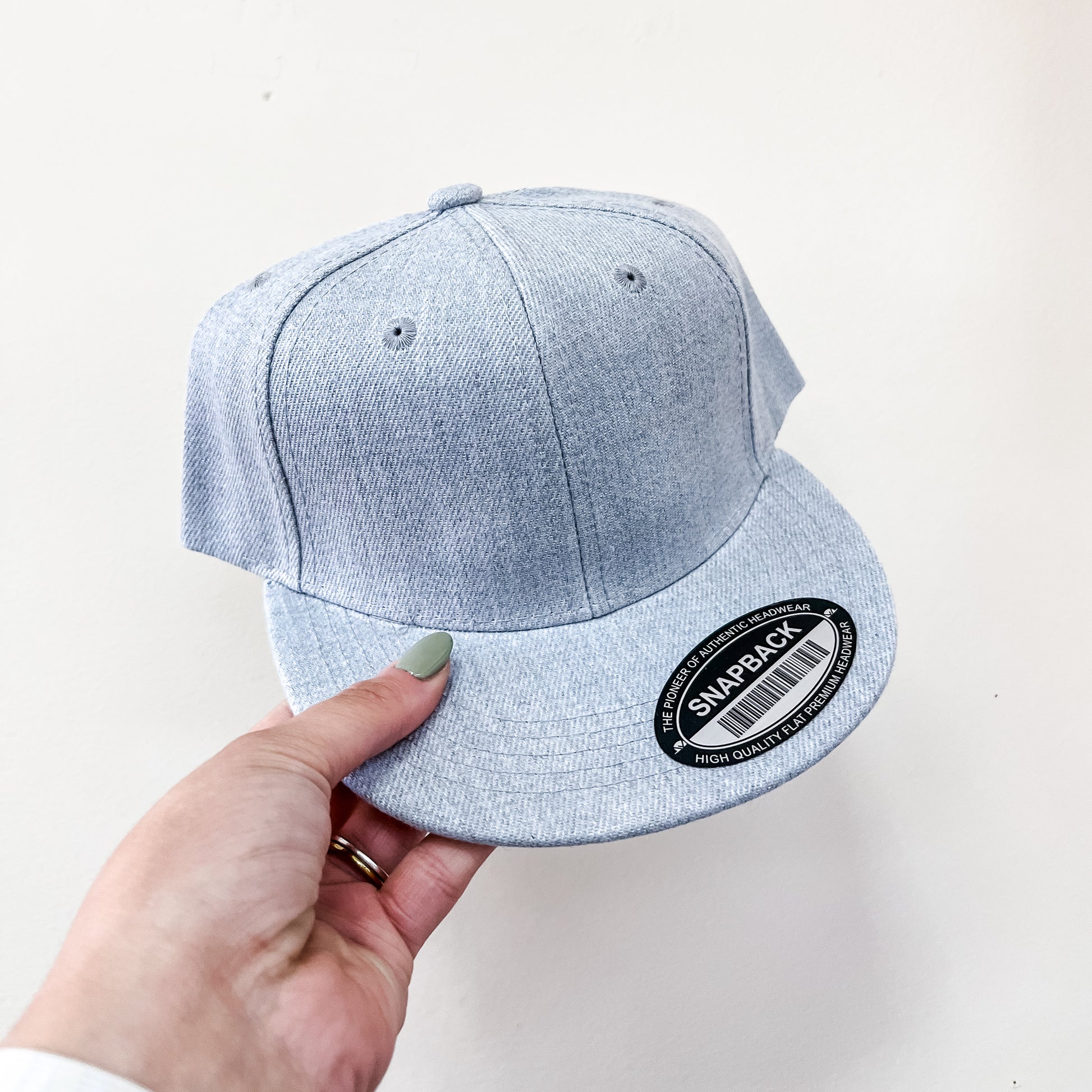 A hand with light green nail polish holds the Capzone Light Grey Snap Back Cap, featuring a round black sticker labeled "SNAPBACK" on the brim. The background is plain white.