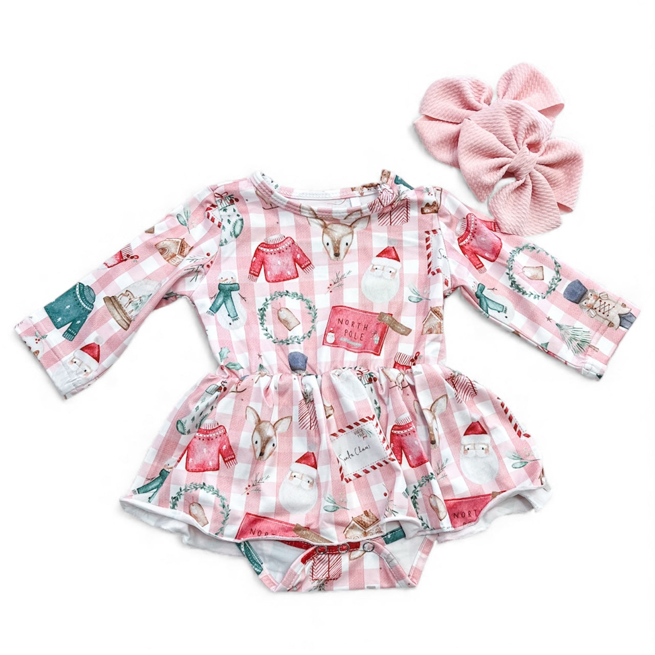 The North Pole Cozy Longsleeve Twirl Romper by Tiny Tot Bundles is a pink and white plaid baby romper with festive holiday prints. Pink textured bow hair clips are placed beside the romper.