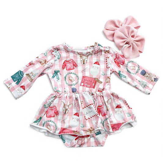 The North Pole Cozy Longsleeve Twirl Romper by Tiny Tot Bundles is a pink and white plaid baby romper with festive holiday prints. Pink textured bow hair clips are placed beside the romper.