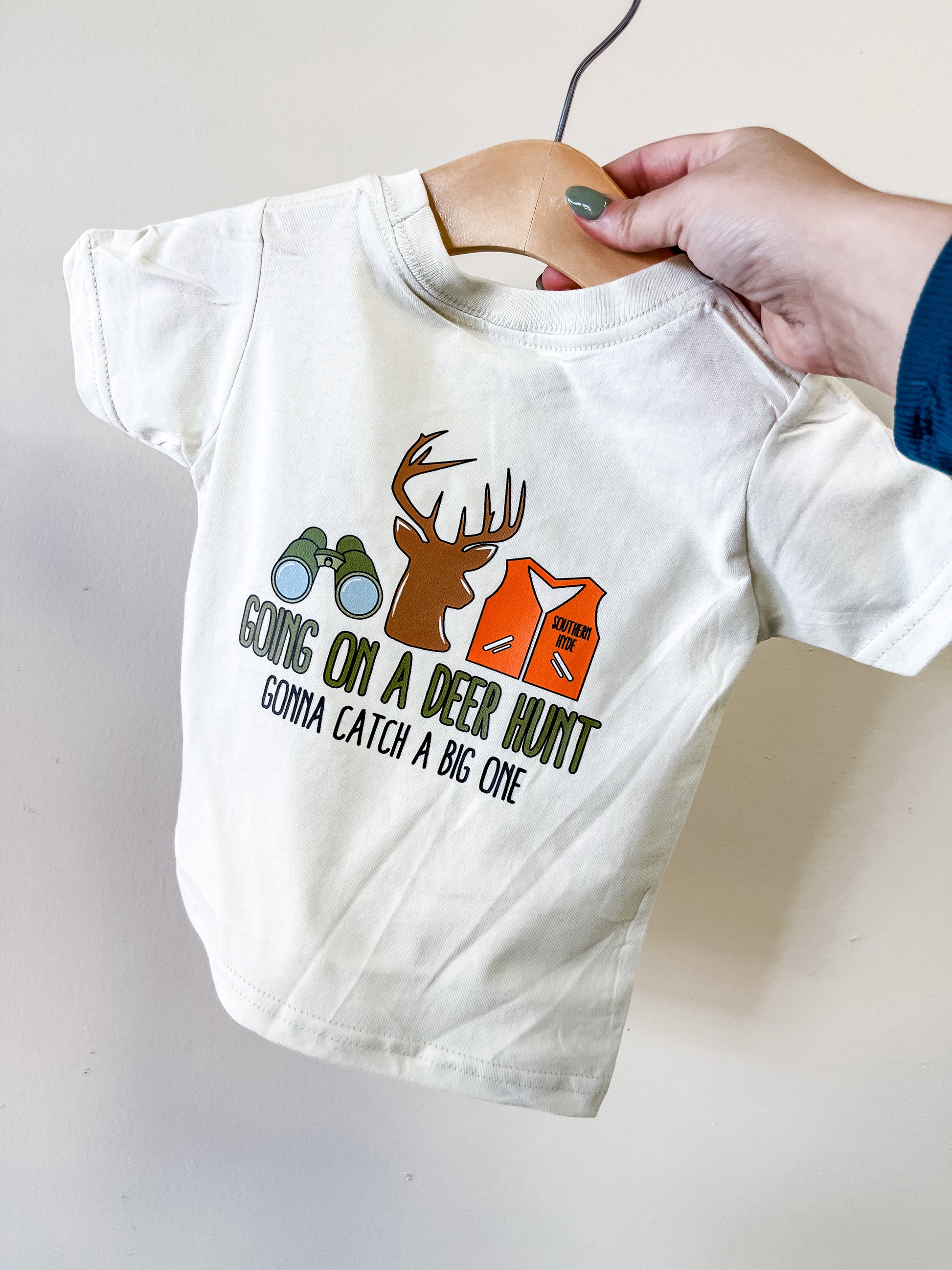 A hand holds up the Southern Hyde Co "Going On A Deer Hunt" T-shirt—a child’s white tee featuring binoculars, a deer head, and an orange vest graphic with the text: Going on a deer hunt, gonna catch a big one.