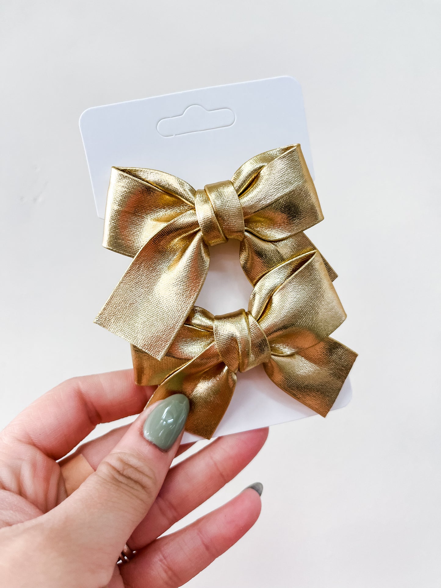 A hand with green-painted nails holds a white card featuring the Shimmer Bow Duo by Tiny Tot Bundles—two shiny gold hair bows—against a plain light background.