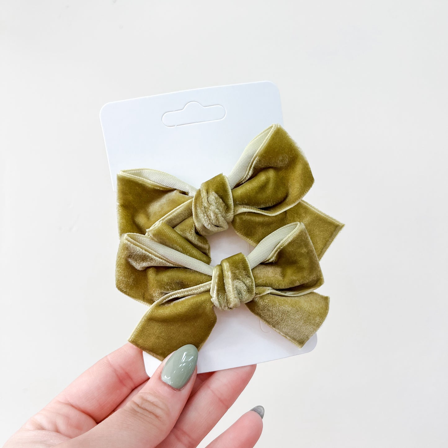 A hand with green-painted nails holds a white card showcasing the Velvet Charm Bow Duo by Tiny Tot Bundles—two matching olive green velvet hair bows—set against a plain light background.