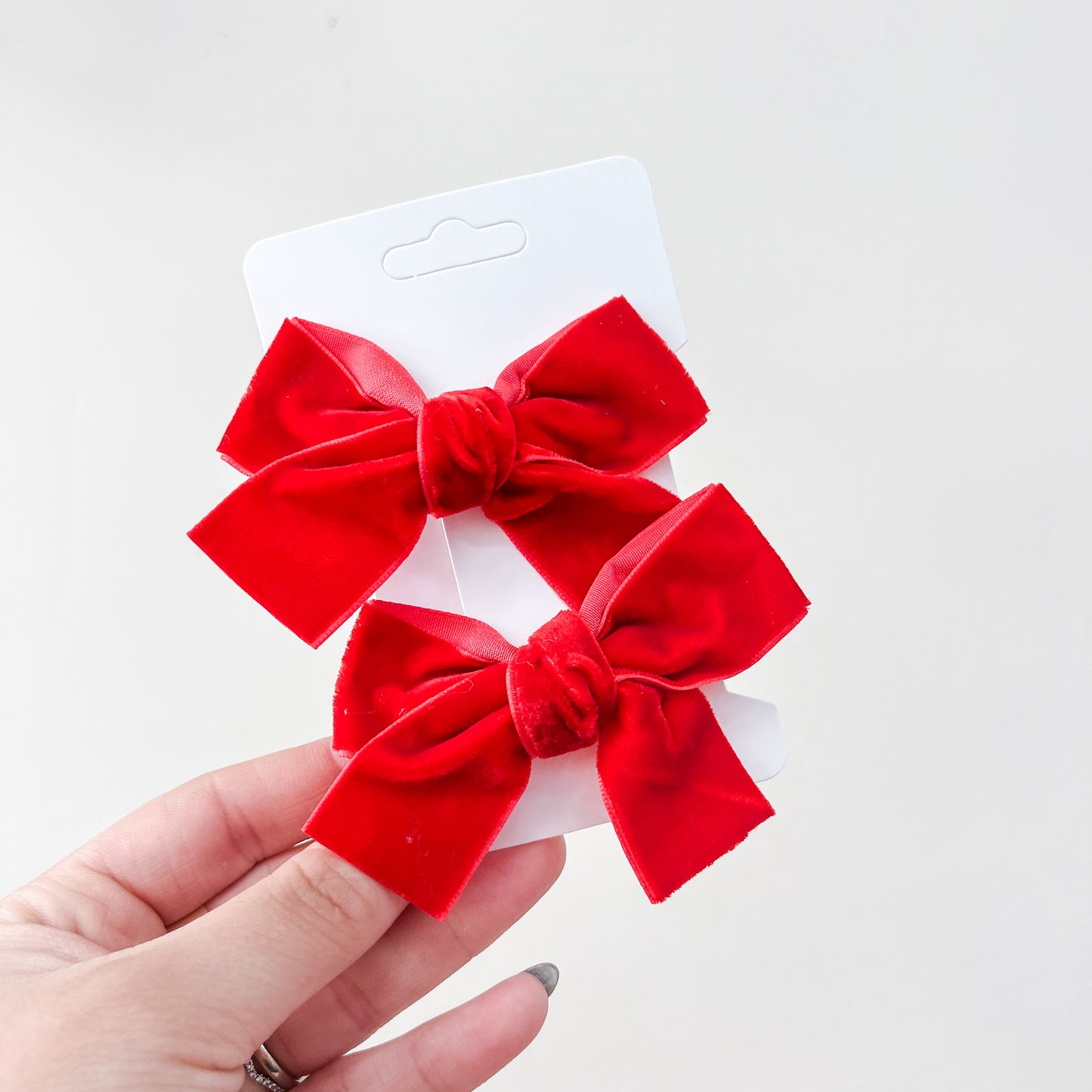 A hand displays Tiny Tot Bundles' Velvet Charm Bow Duo—two bright red velvet hair bows in classic shapes—set against a plain white background.