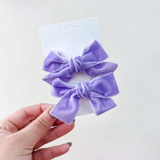 A hand holds a white card featuring Tiny Tot Bundles' Velvet Charm Bow Duo—two purple velvet hair bows with knot centers—set against a plain light background.