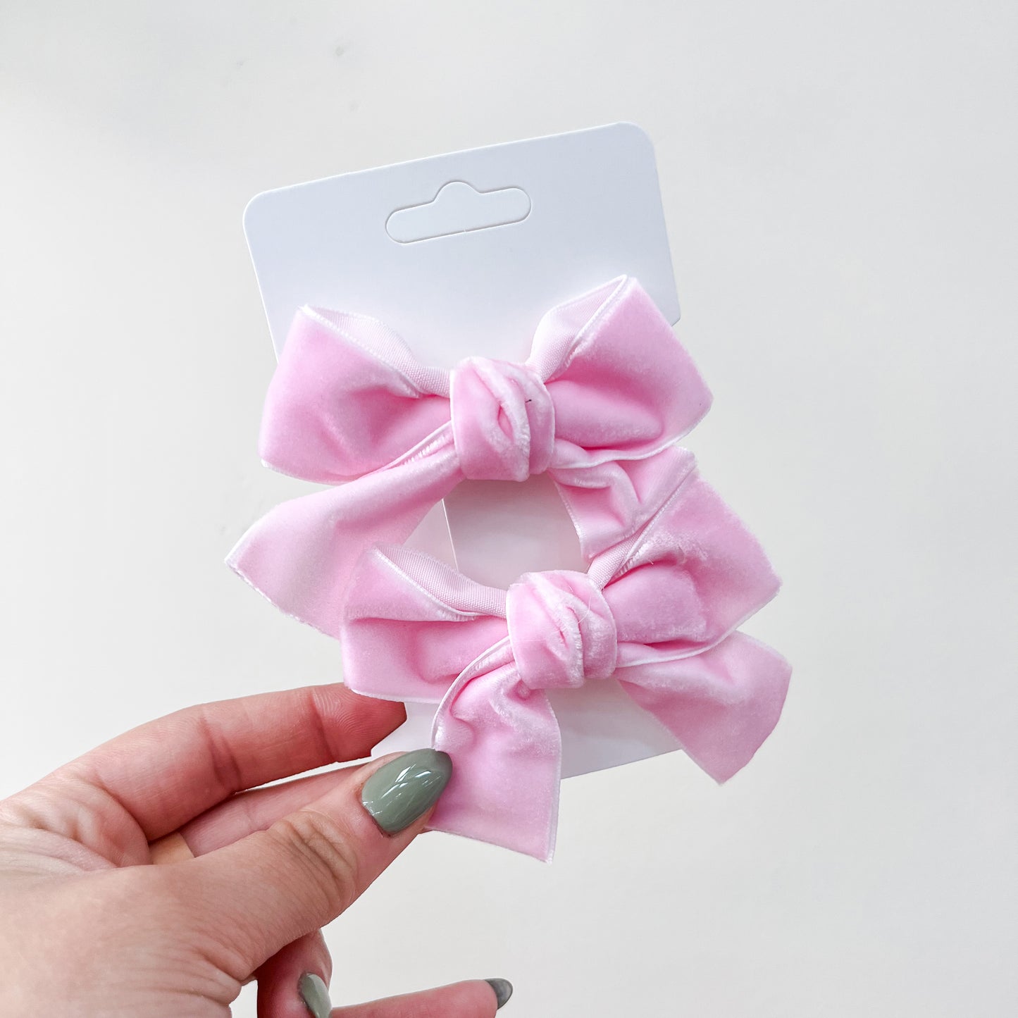 A hand with green-painted nails holds a white card displaying the Velvet Charm Bow Duo by Tiny Tot Bundles—two light pink velvet hair bows—against a plain white background.
