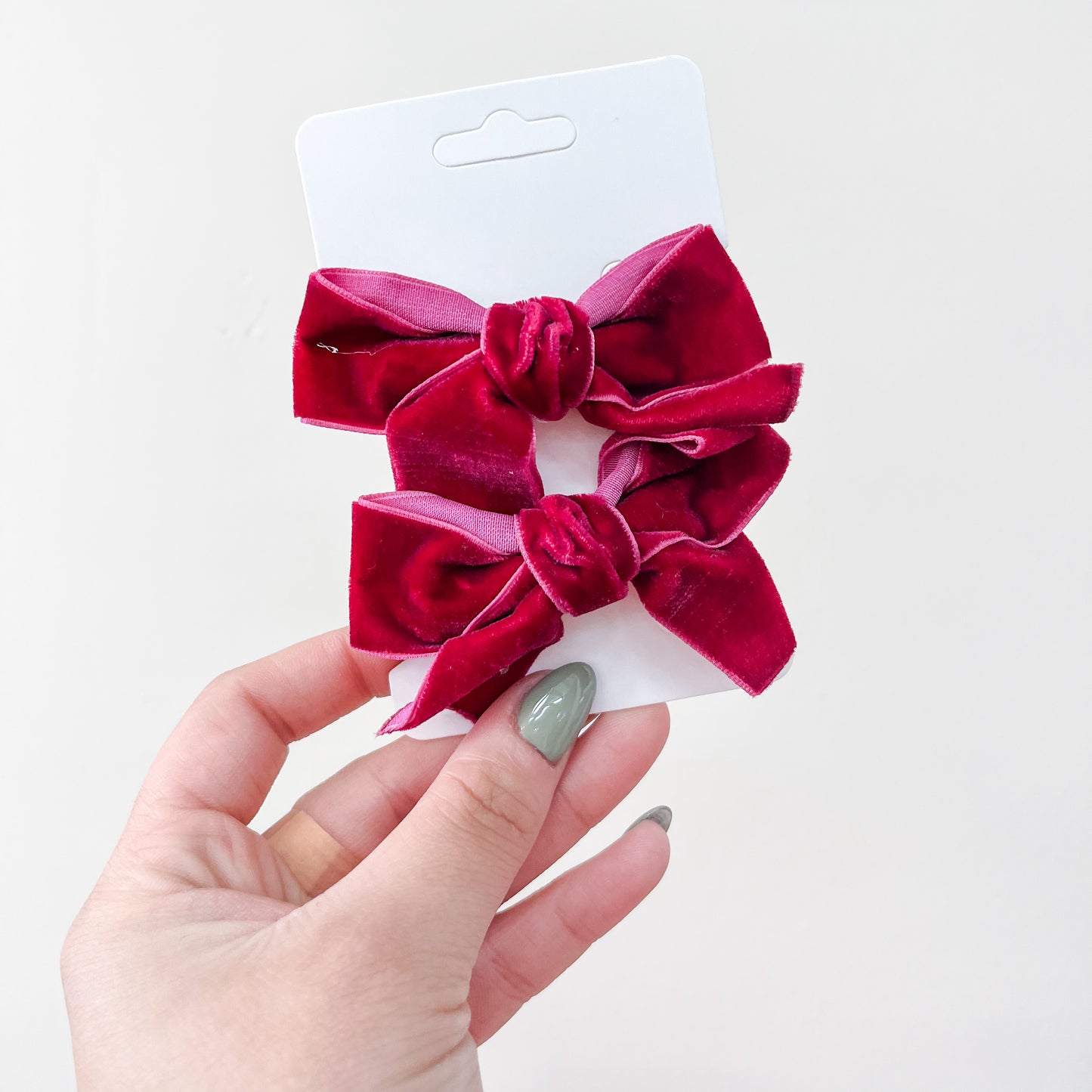 A hand with painted nails holds a white card featuring the Velvet Charm Bow Duo by Tiny Tot Bundles—two dark pink velvet hair bows with central knots—set against a plain light background.