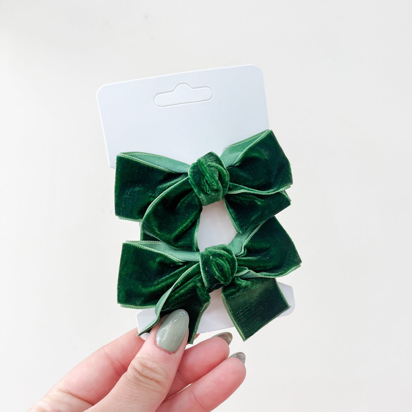 A hand with green-painted nails holds a white card displaying the Velvet Charm Bow Duo by Tiny Tot Bundles—two matching dark green velvet hair bows—against a plain light background.