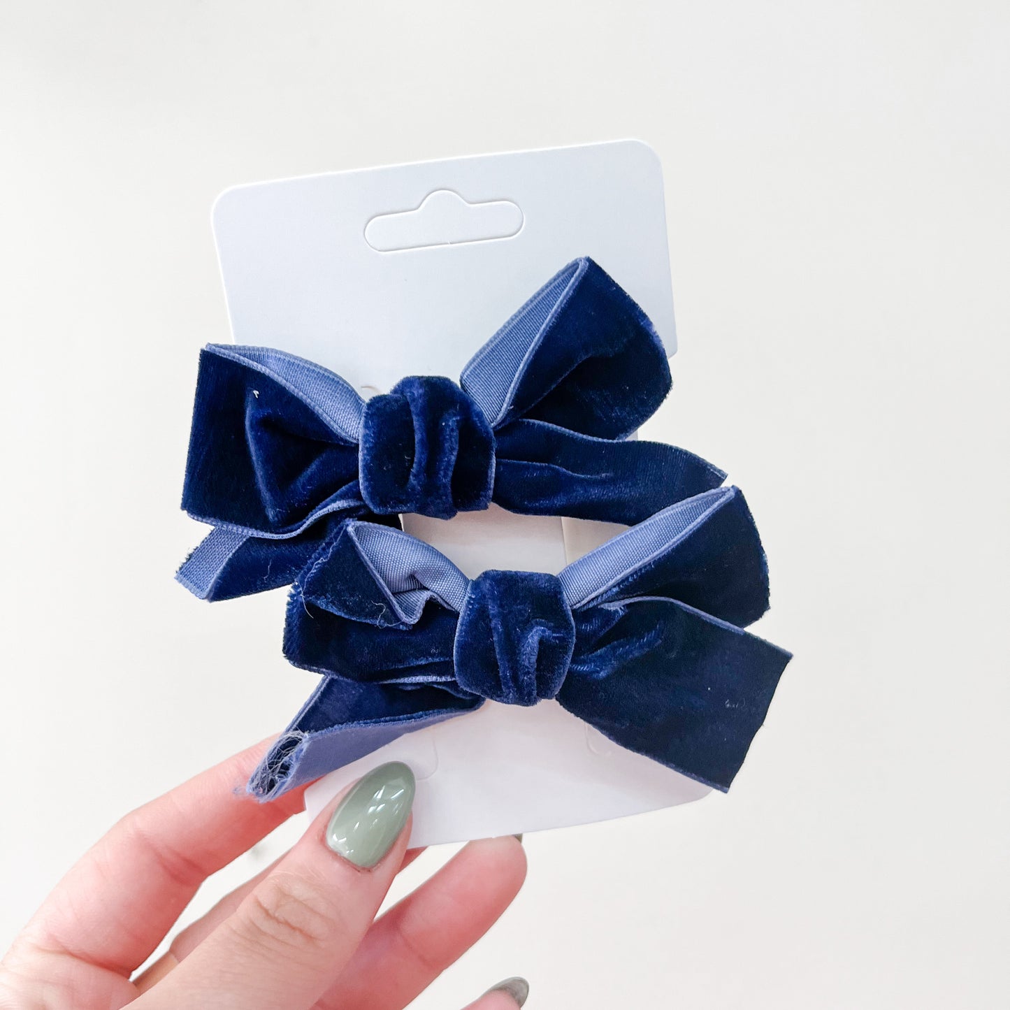 A hand with painted nails holds a white card featuring the Velvet Charm Bow Duo by Tiny Tot Bundles—two dark blue velvet hair bows with knotted centers—against a plain light background.