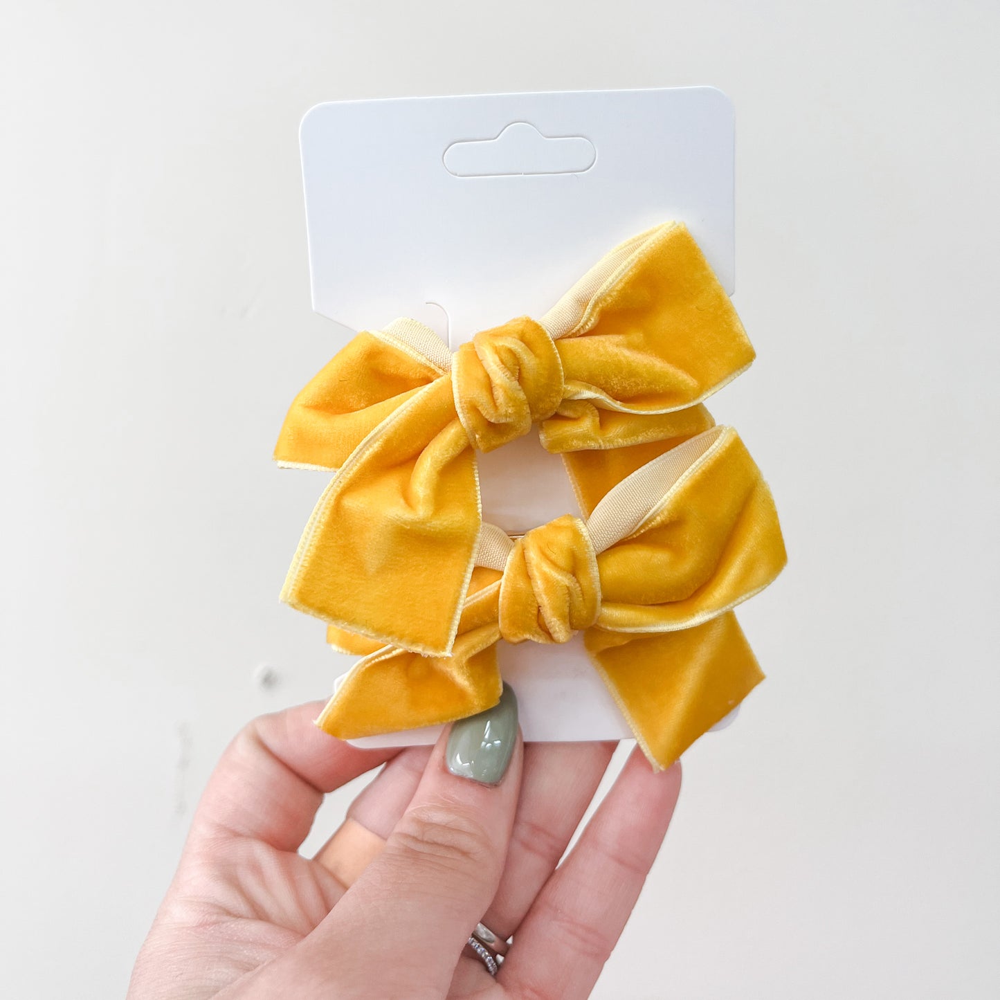 A hand with light green nail polish holds a white card displaying the Velvet Charm Bow Duo by Tiny Tot Bundles—two bright yellow velvet bows, each knotted in the center—against a plain light background.
