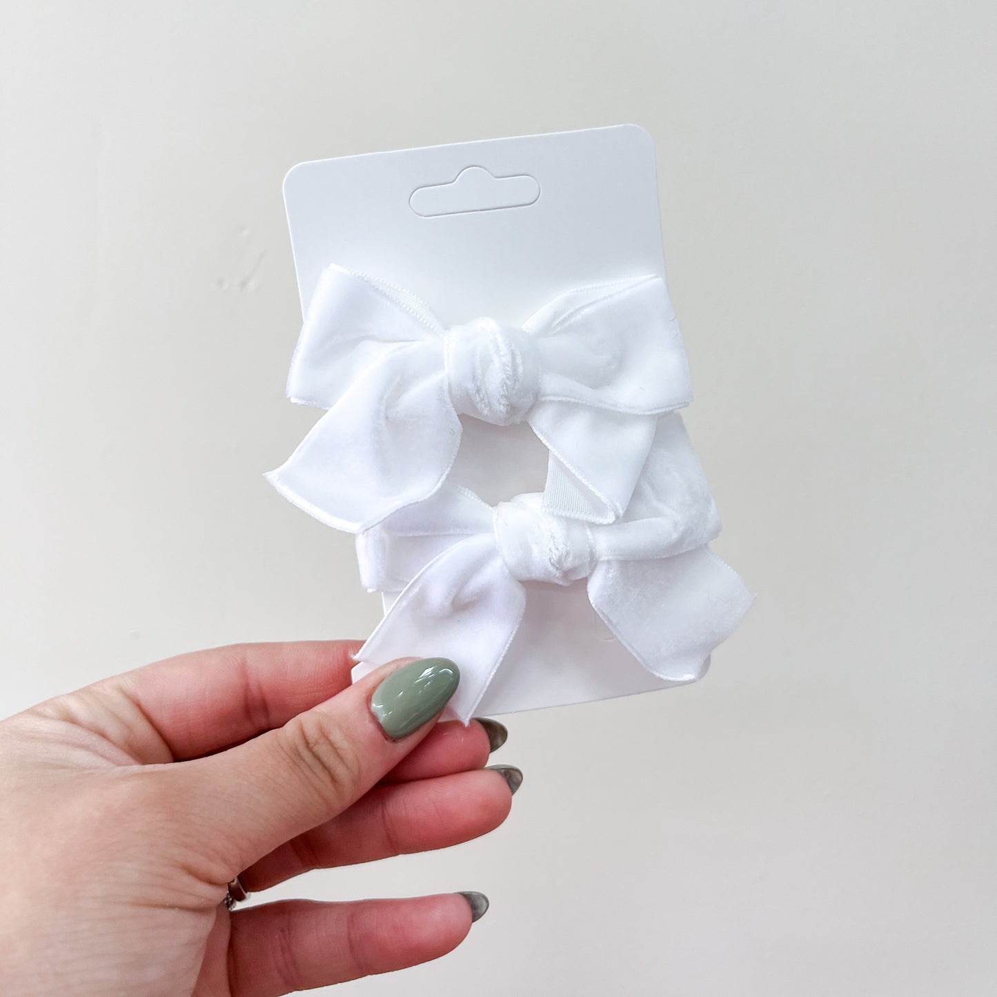 A hand with green-painted nails holds a Tiny Tot Bundles Velvet Charm Bow Duo on a white cardboard backing, displaying two white fabric bows against a plain light background.