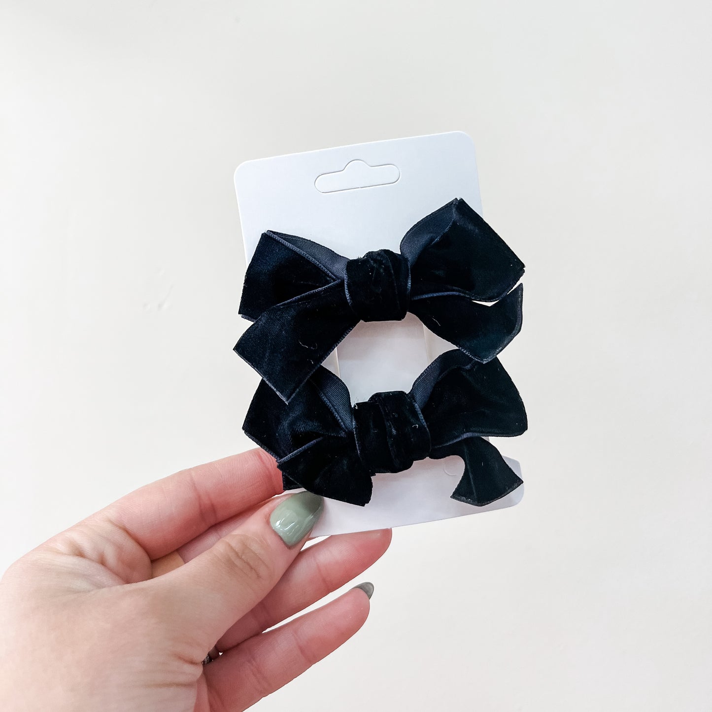 A hand with painted nails holds a white card displaying the Tiny Tot Bundles Velvet Charm Bow Duo—two black velvet hair bows—against a plain light background.