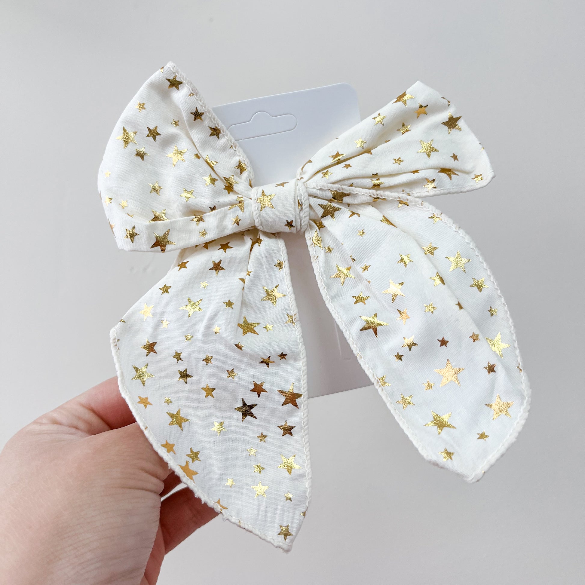 A hand holds the Tiny Tot Bundles Holiday Cheer Bow Set featuring a white fabric bow with shiny gold stars, displayed on its card against a plain light background.