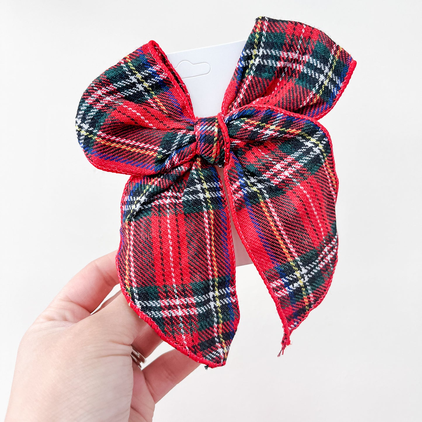 A hand displays a large Holiday Cheer Bow from Tiny Tot Bundles, featuring red plaid fabric with green, yellow, white, and black stripes and two long tails, set against a plain white background.