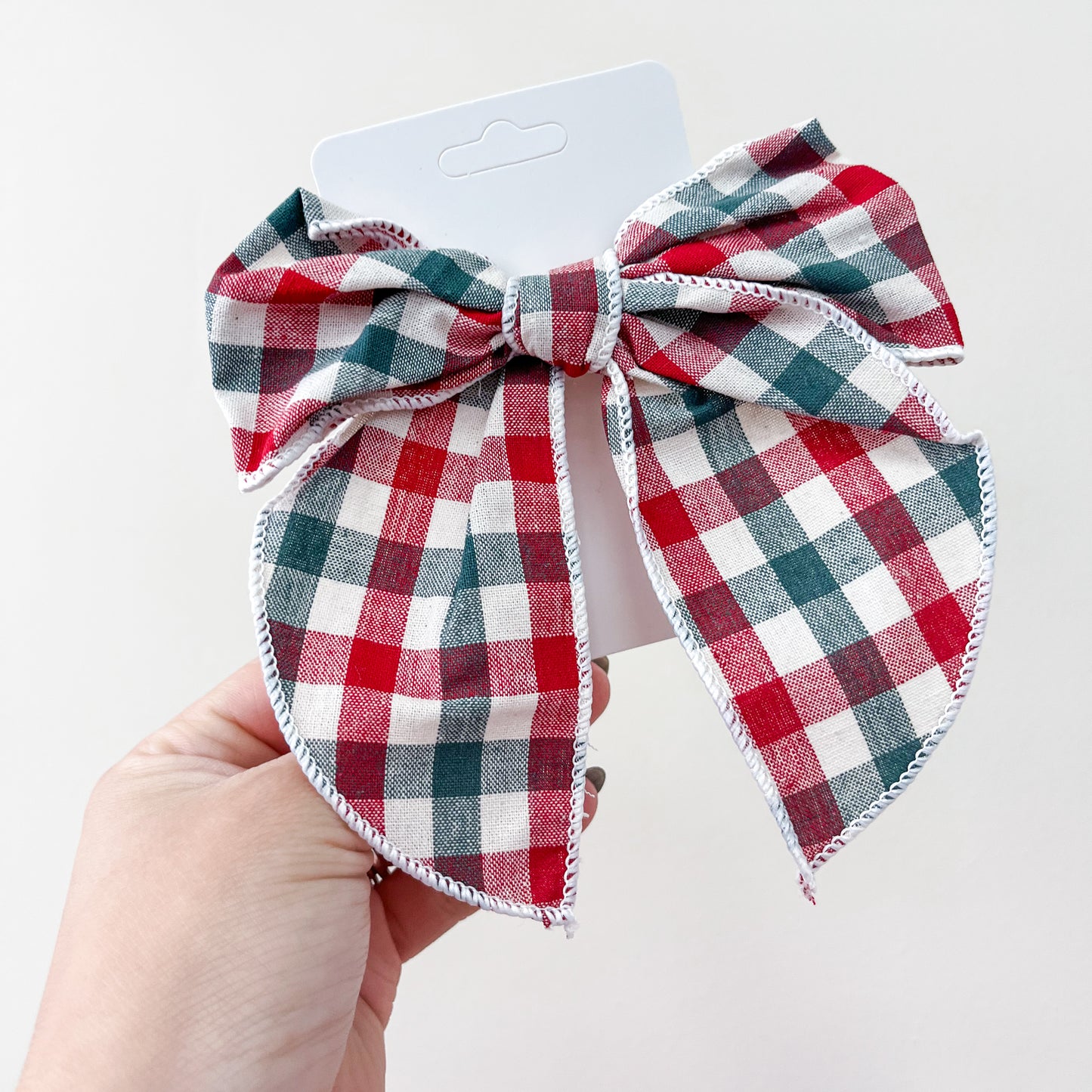 A hand holds the Holiday Cheer Bow Set by Tiny Tot Bundles—a large red, green, and white plaid bow with long, white-stitched tails, attached to a white card with a hanger slot.