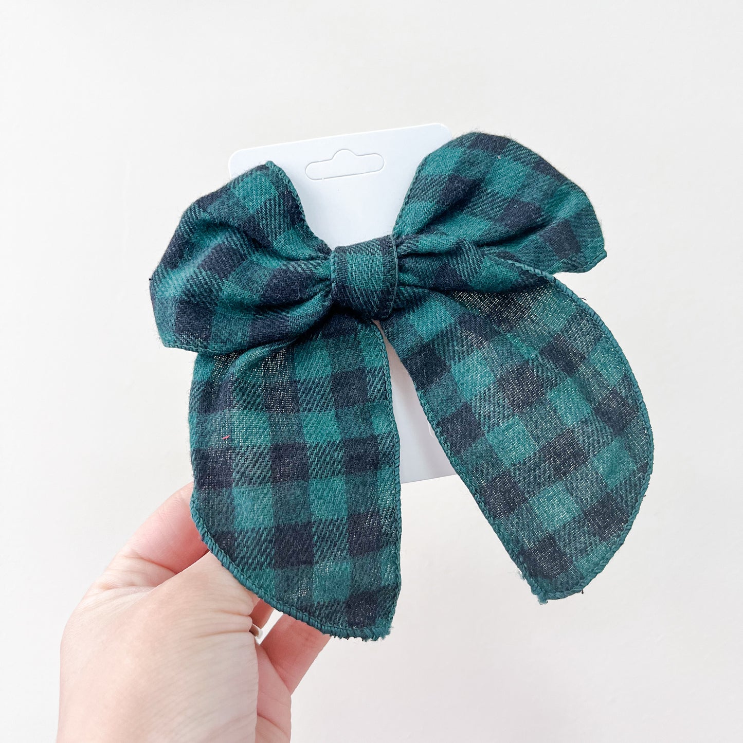 A hand holds a black and green plaid fabric bow from the Holiday Cheer Bow Set by Tiny Tot Bundles, attached to a white display card against a plain white background.
