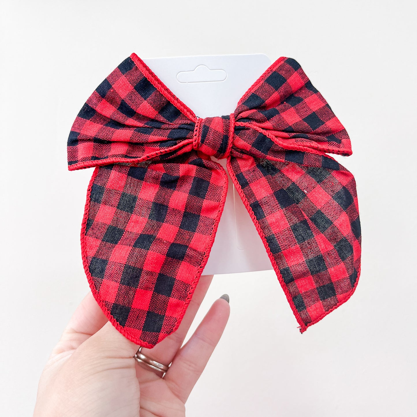 A hand holds a red and black buffalo plaid bow from the Holiday Cheer Bow Set by Tiny Tot Bundles, attached to a white card against a plain white background.