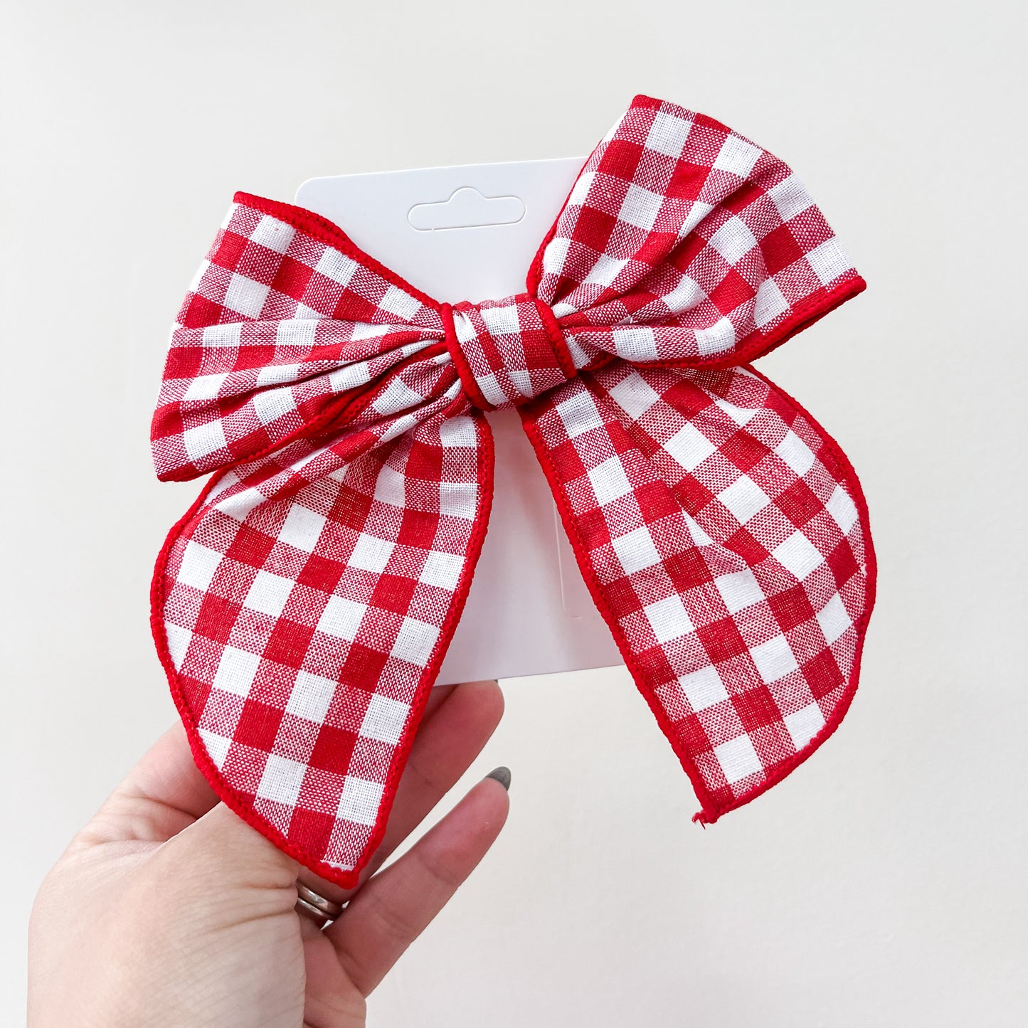 A hand holds a large red and white checkered bow from the Tiny Tot Bundles Holiday Cheer Bow Set, attached to a white card and displayed against a plain light background.