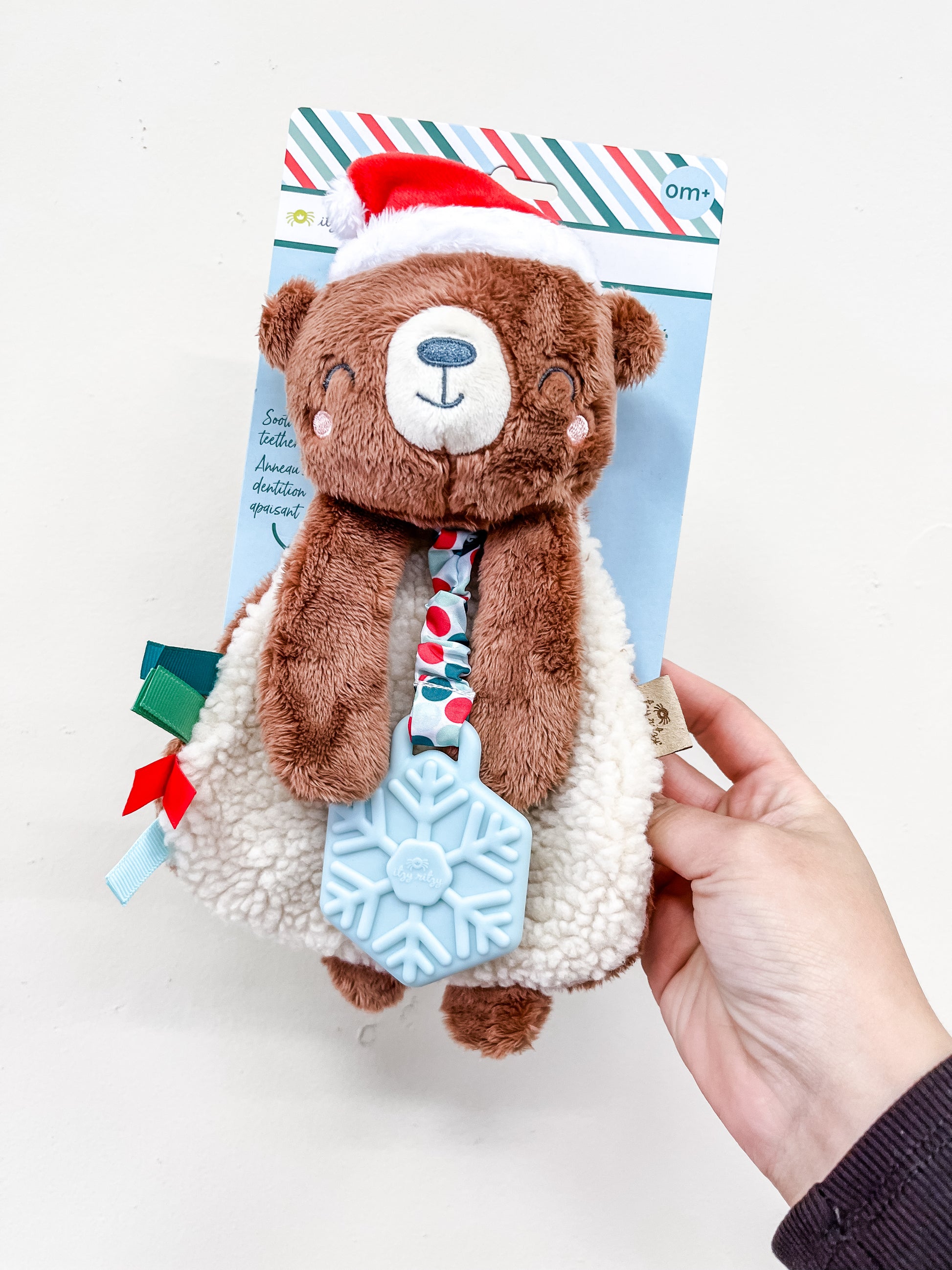 A hand holds the itzy ritzy Holiday Bear Plush + Teether, featuring a soft white bear with a Santa hat, festive tie, and a blue snowflake-shaped teething toy attached to its hand.