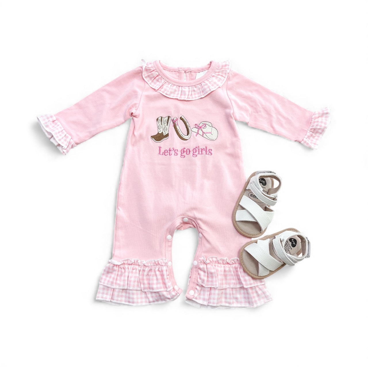 The Tiny Tot Bundles Let’s Go Girls Romper, featuring a pink design with ruffled collar and cuffs, boots and horseshoe graphics, is paired with white baby sandals and shown on a white background.