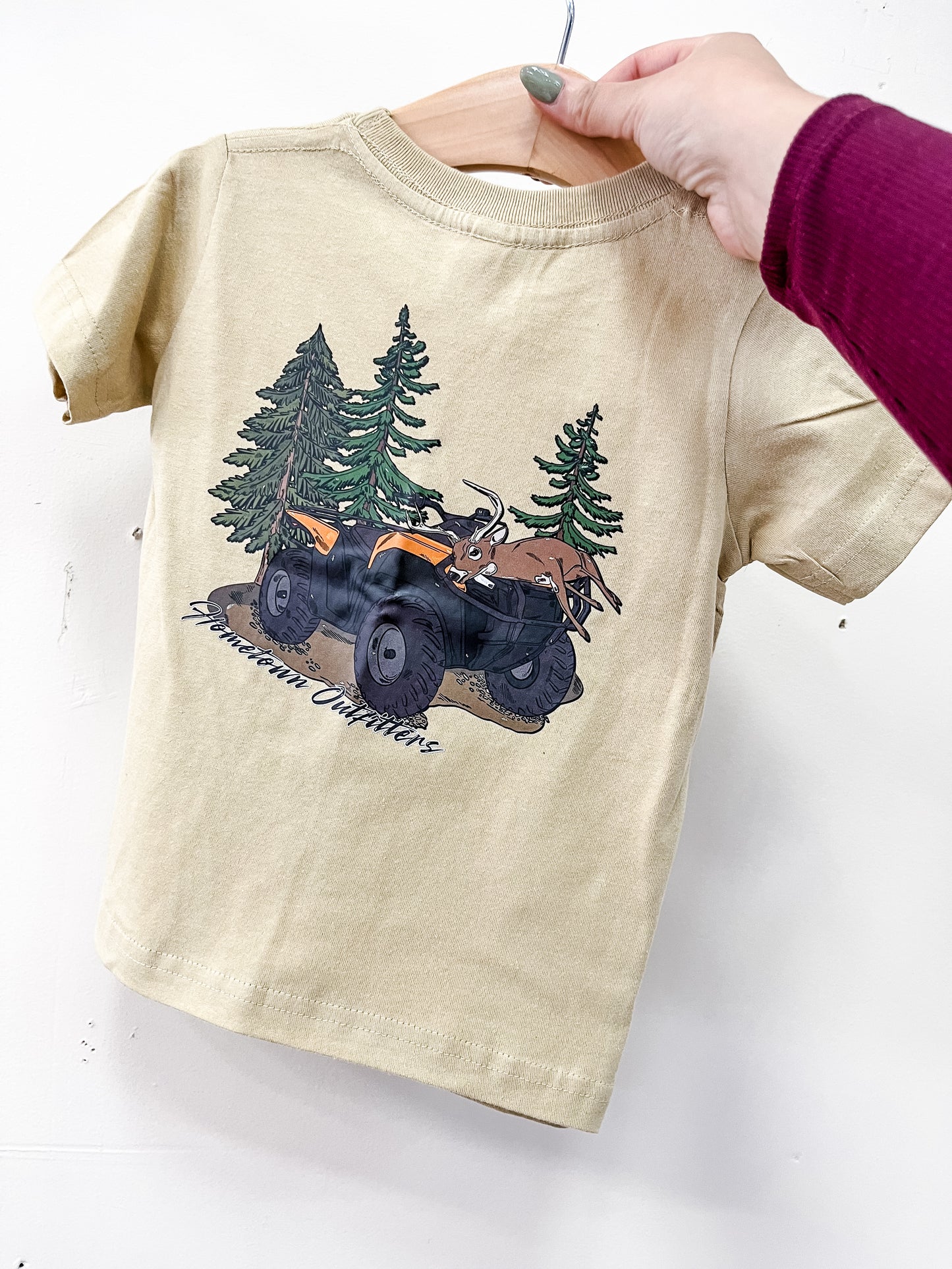 A beige Folded and Loaded T-Shirt by hometown Outfitters hangs on a wooden hanger held by a hand. It features an ATV and pine trees graphic with "Mountain Outfitters" text beneath the illustration.