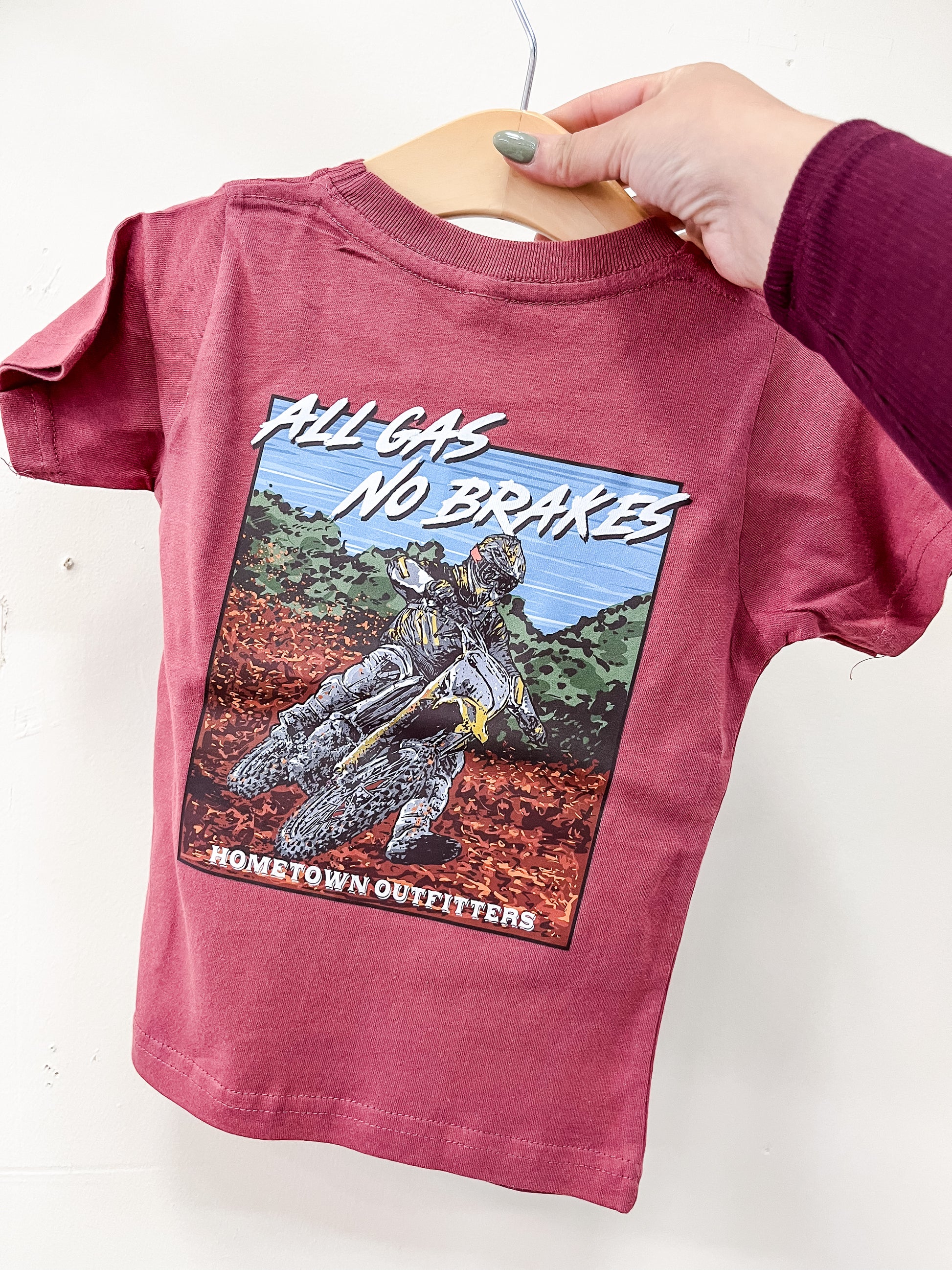 A hand holds a red All Gas No Brakes T-Shirt by hometown Outfitters, featuring a motocross rider graphic with the text "ALL GAS NO BRAKES" and "hometown Outfitters.
