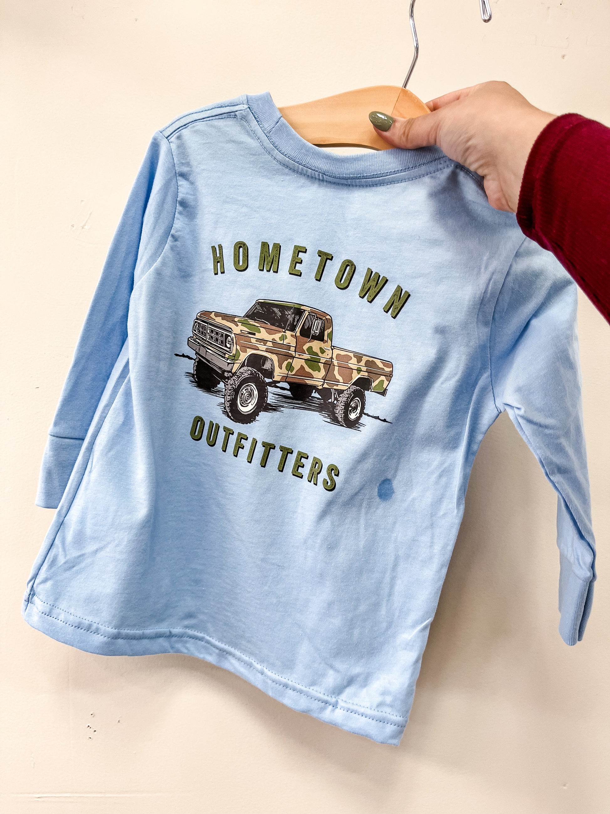 A hand with painted nails holds the Old School Long Sleeve T-shirt by hometown Outfitters—a light blue children’s shirt featuring a camouflage truck graphic and “HOMETOWN OUTFITTERS”—on a wooden hanger against a beige background.