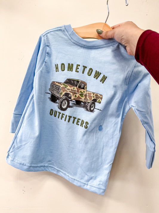 A hand with painted nails holds the Old School Long Sleeve T-shirt by hometown Outfitters—a light blue children’s shirt featuring a camouflage truck graphic and “HOMETOWN OUTFITTERS”—on a wooden hanger against a beige background.