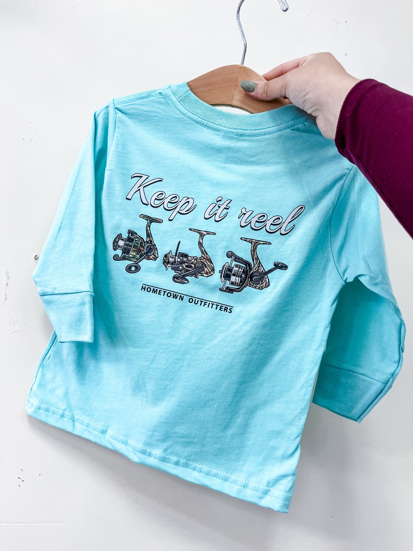 A person holds a light blue Keep It Reel Long Sleeve by hometown Outfitters on a hanger. The back displays fishing reel drawings with "Keep it reel" above "hometown Outfitters.