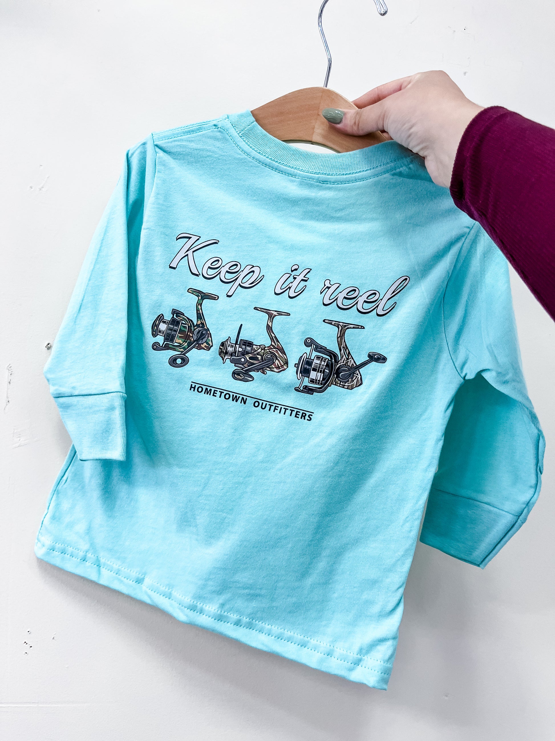 A person holds a light blue Keep It Reel Long Sleeve by hometown Outfitters on a hanger. The back displays fishing reel drawings with "Keep it reel" above "hometown Outfitters.