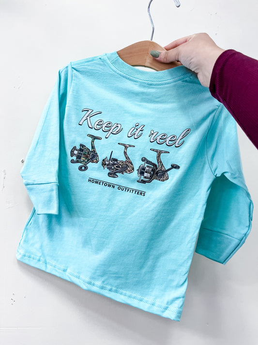 A person holds a light blue Keep It Reel Long Sleeve by hometown Outfitters on a hanger. The back displays fishing reel drawings with "Keep it reel" above "hometown Outfitters.