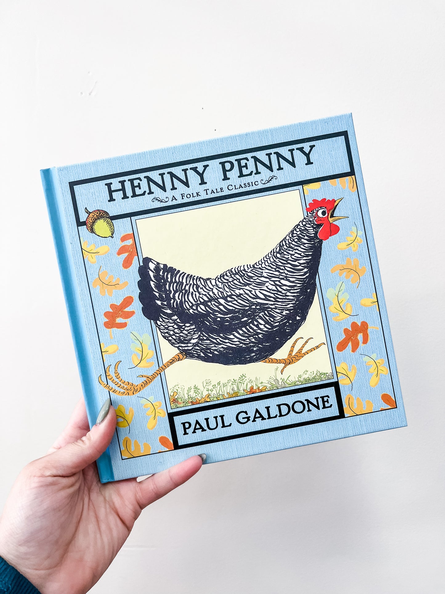 A hand holds the Henny Penny Book by HarperCollins, featuring a black hen with a red comb amid yellow and orange leaves on a light blue cover.