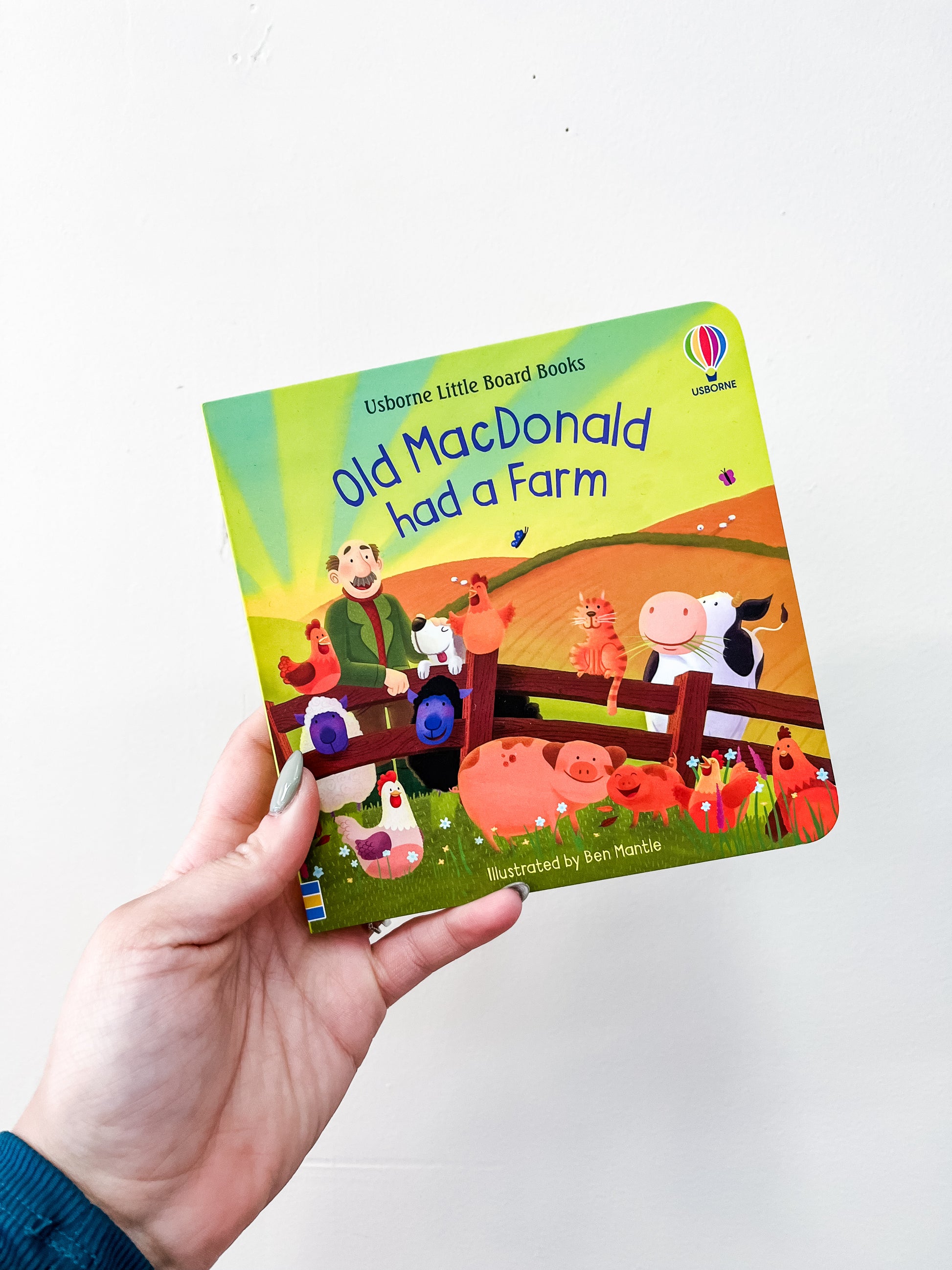 A hand holds the HarperCollins Old Macdonald Had A Farm Board Book, which features colorful cover illustrations of a farmer, farm animals, and a barn.