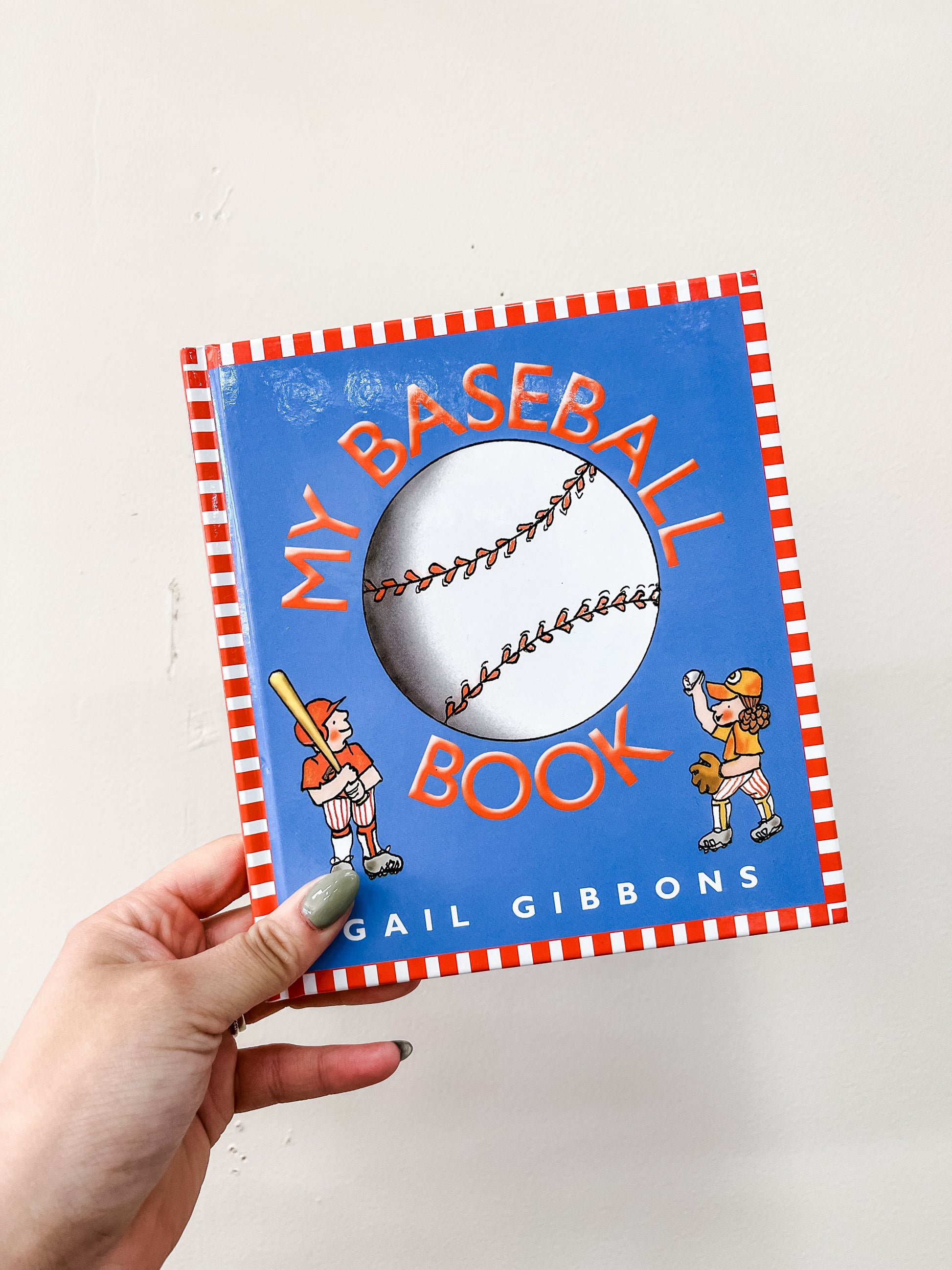 A hand holds the HarperCollins children’s book "My Baseball Book," which has a blue cover featuring a large baseball and cartoon kids in uniforms.
