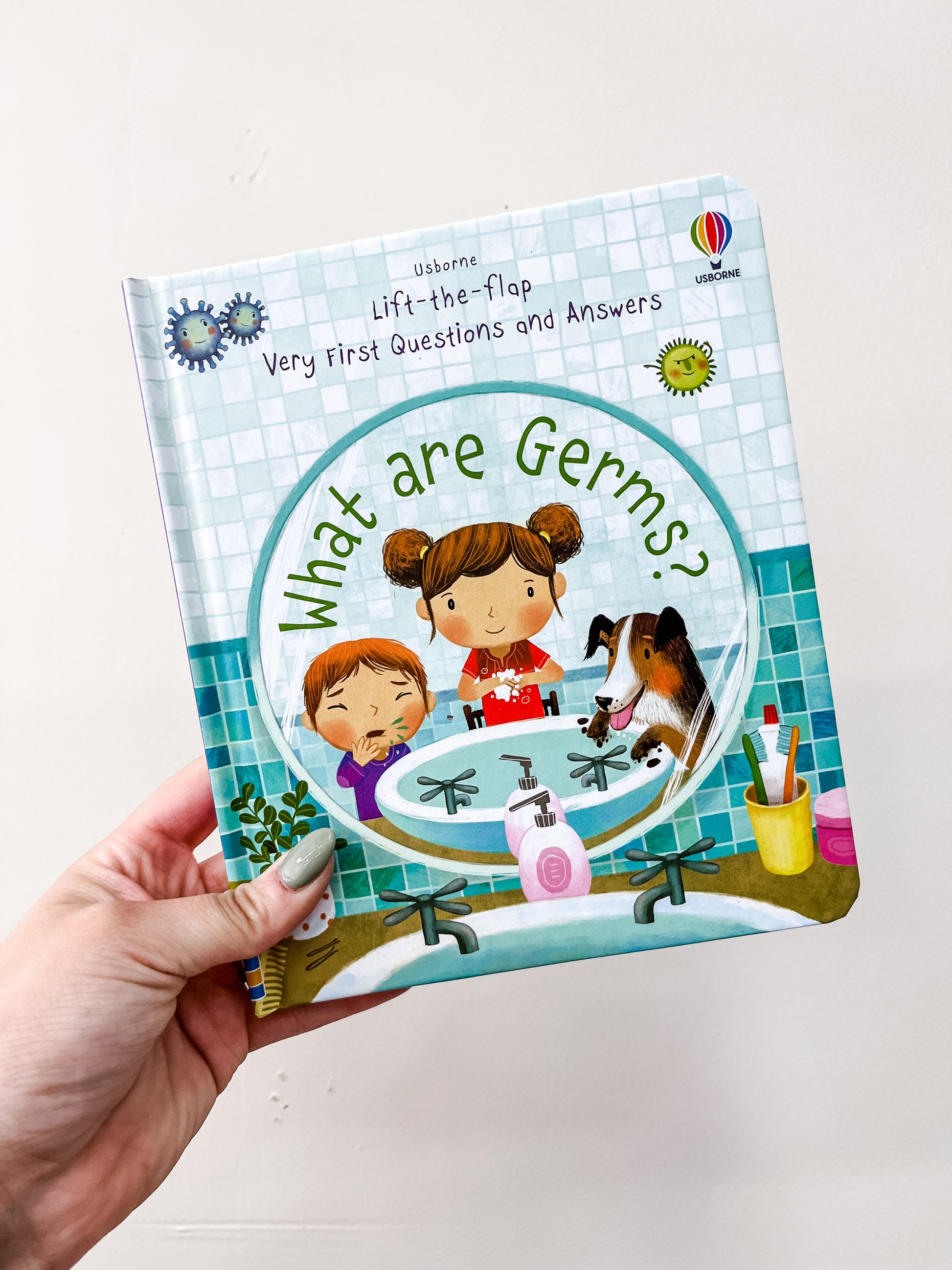 A hand holds the "What Are Germs Book" by HarperCollins, featuring cartoon kids and a dog washing hands at a sink, with illustrated germs above the title.