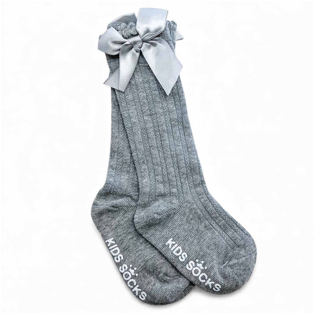Tiny Tot Bundles Pretty Bow Ribbed Socks are gray ribbed kids’ socks with “KIDS SOCKS” in white on the soles and a large silver satin bow at the top, shown laid flat against a white background.