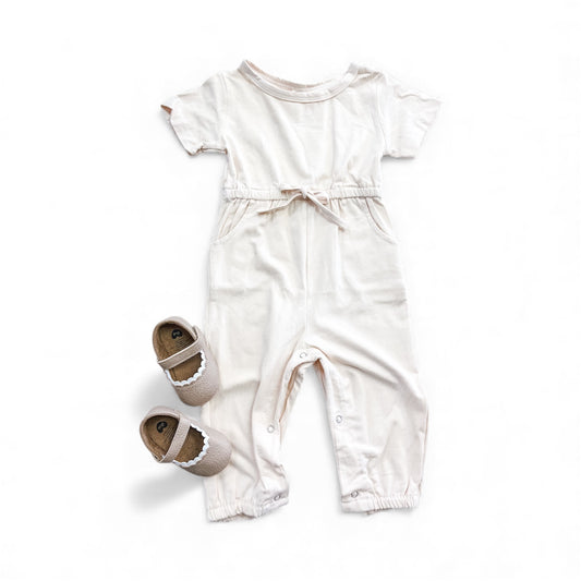 The Sweet Simplicity Jumpsuit by Tiny Tot Bundles, a light pink short-sleeve romper with a waist tie and snap buttons, is shown beside beige scalloped baby shoes on a white background.