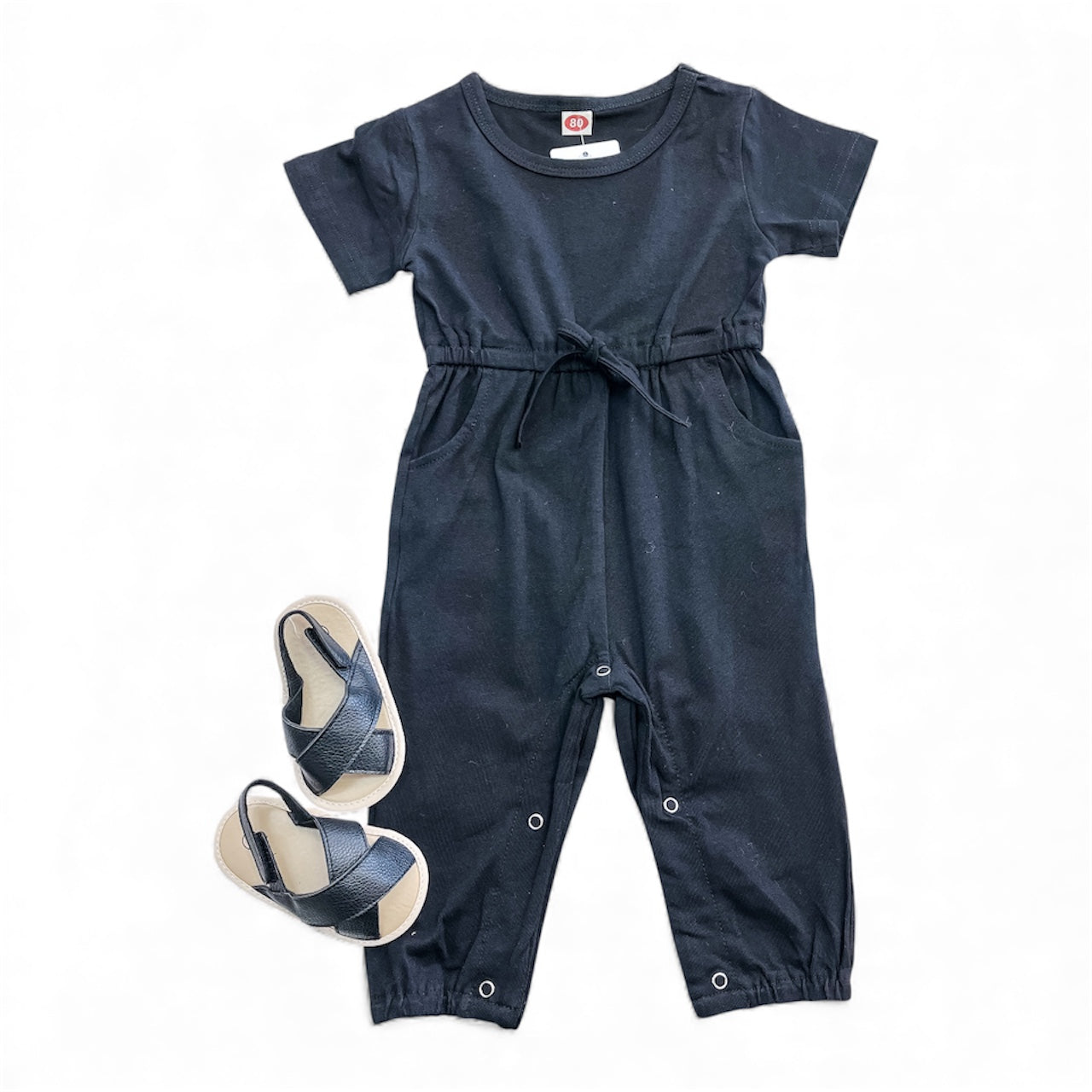 The Sweet Simplicity Jumpsuit by Tiny Tot Bundles, a navy blue short-sleeve toddler jumpsuit with a drawstring waist and snap-leg buttons, is paired with matching sandals and shown on a white background.