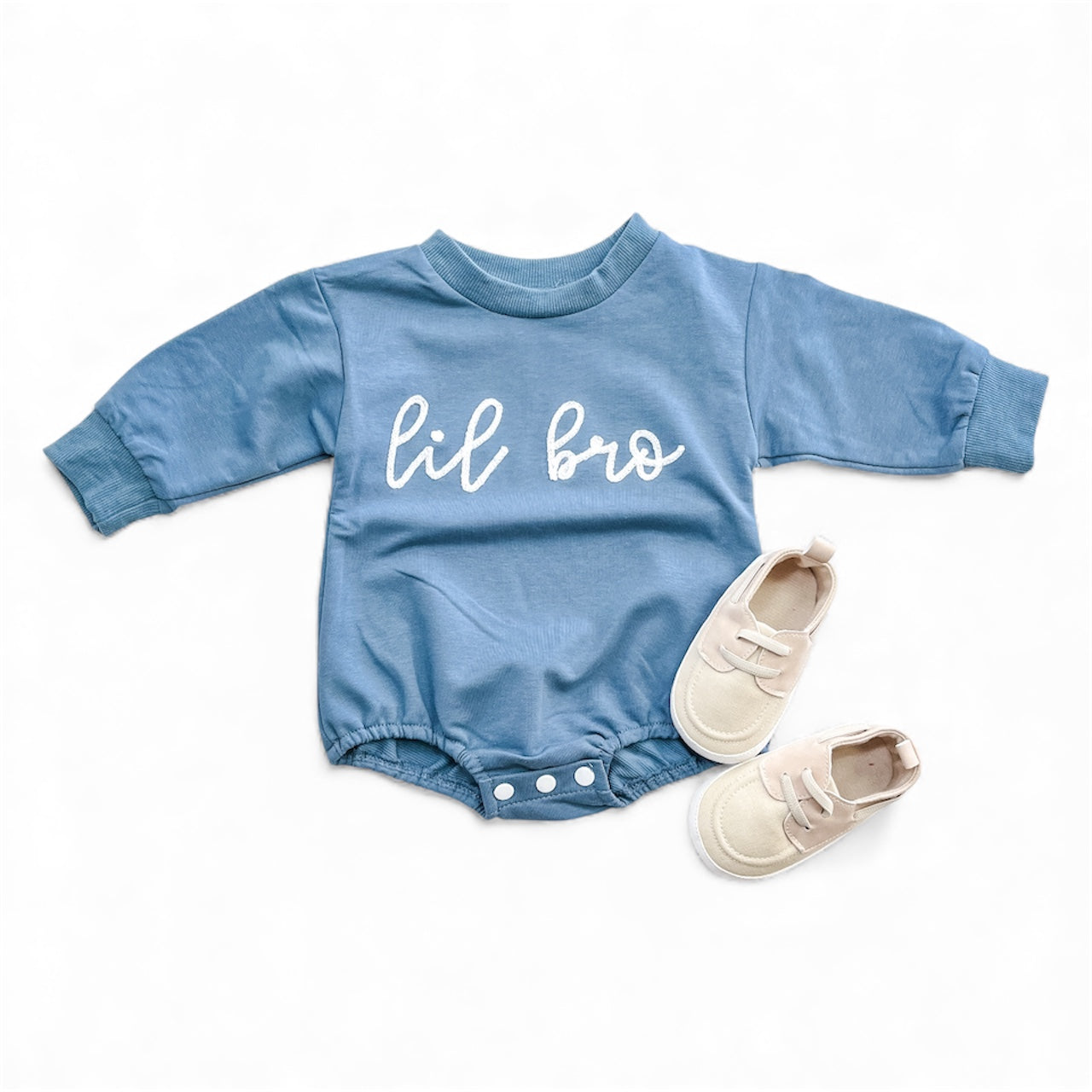 The Tiny Tot Bundles Lil Bro Bubble, a blue long-sleeve romper with "lil bro" in white on the front, is paired with light beige baby shoes and displayed on a white background.