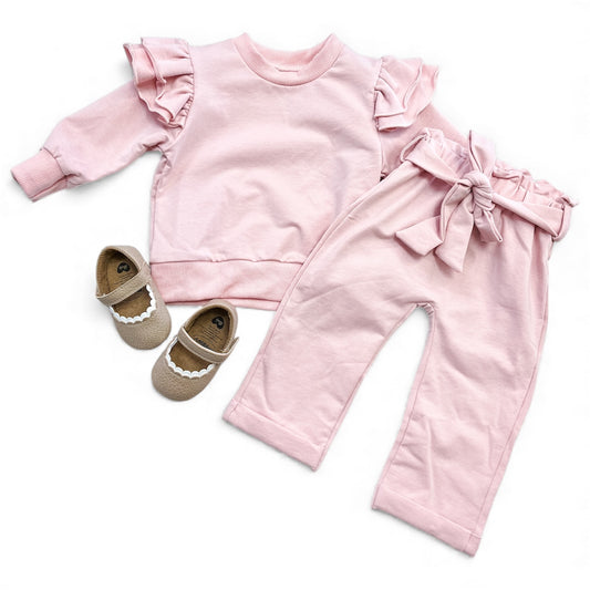 The Blush Breeze Ruffle Set by Tiny Tot Bundles features a light pink ruffled long-sleeve top, matching waist-tied pants, and beige baby shoes with white scalloped trim, all displayed on a white background.
