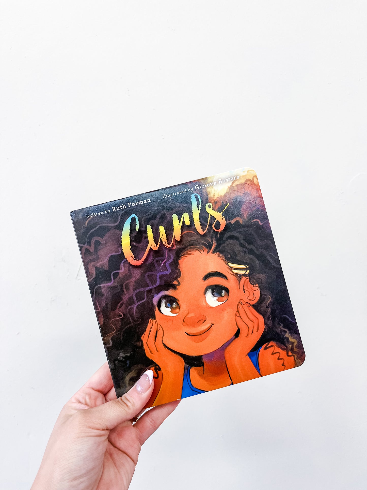 A hand holds the Curls Book from Simon & Schuster, featuring an illustrated girl with curly hair, smiling and looking upward against a plain white background.