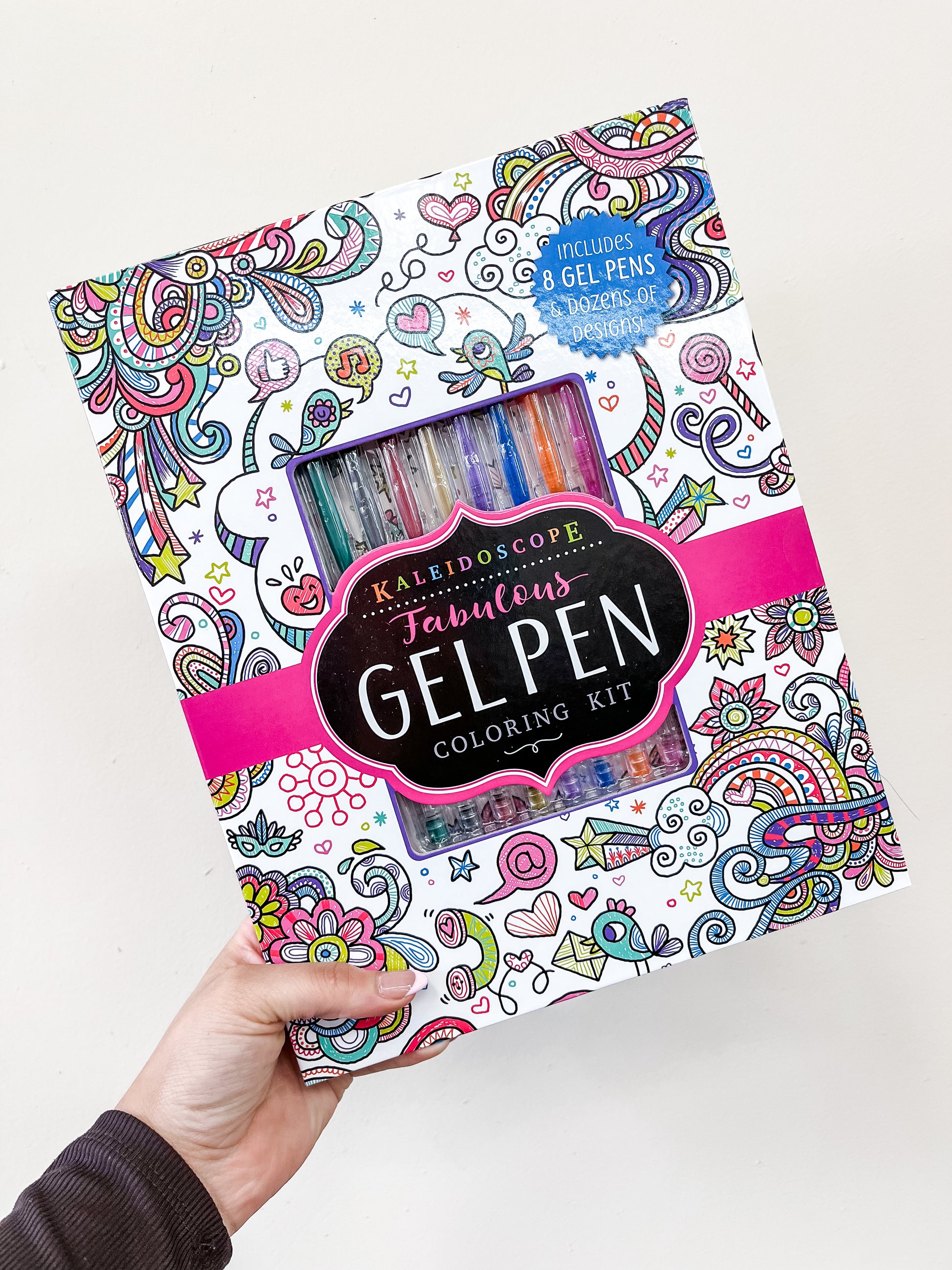 A hand displays the Simon & Schuster Kaleidoscope Fabulous Gel Pen Coloring Kit box with vibrant doodle art, a pink band, and a window revealing 8 gel pens. Text notes the kit contains gel pens and dozens of coloring designs.