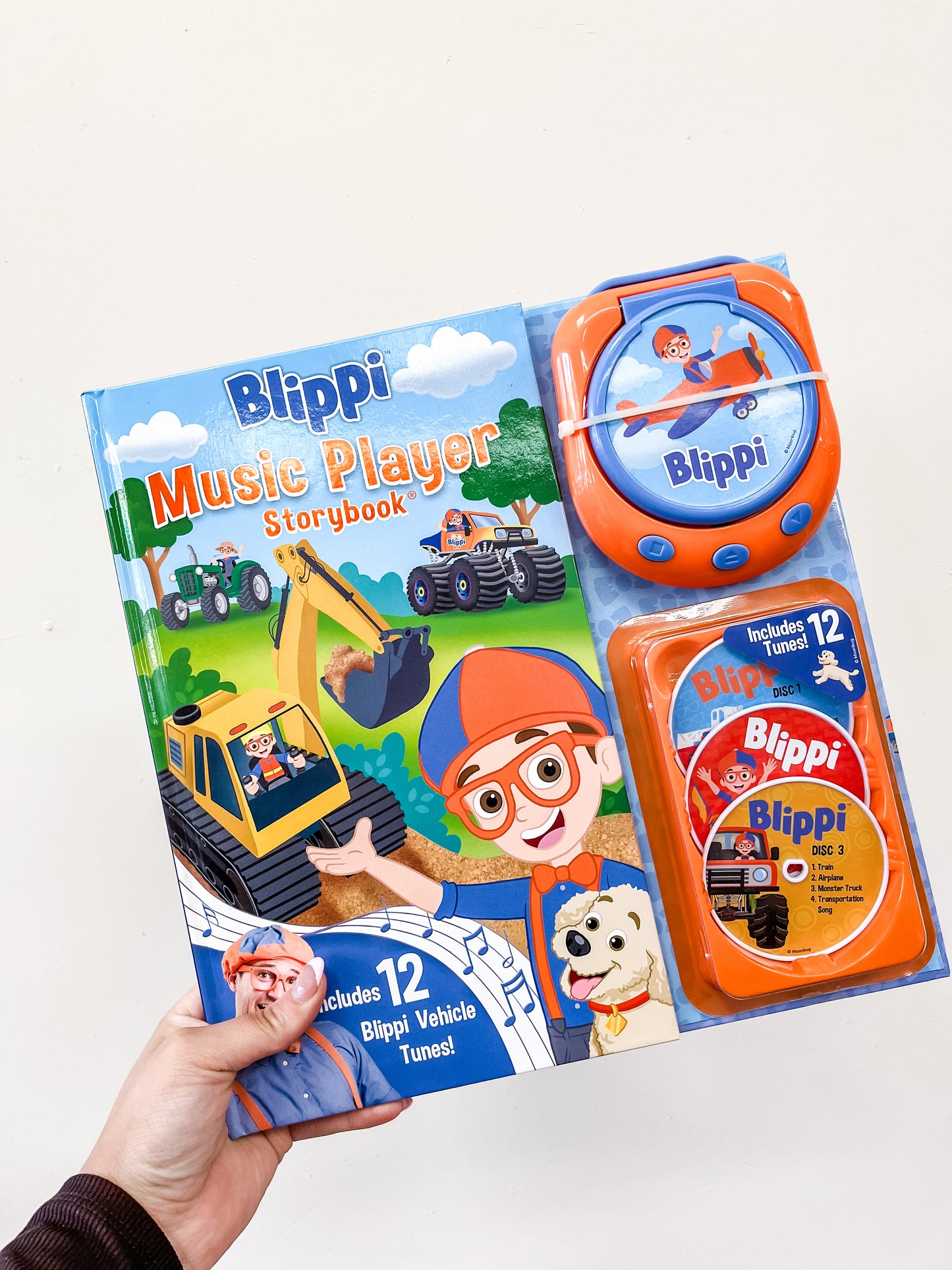 A hand holds the Simon & Schuster Blippi Music Player Storybook, which includes a construction vehicle book, a blue and orange music player, and discs with 12 vehicle-themed tunes.