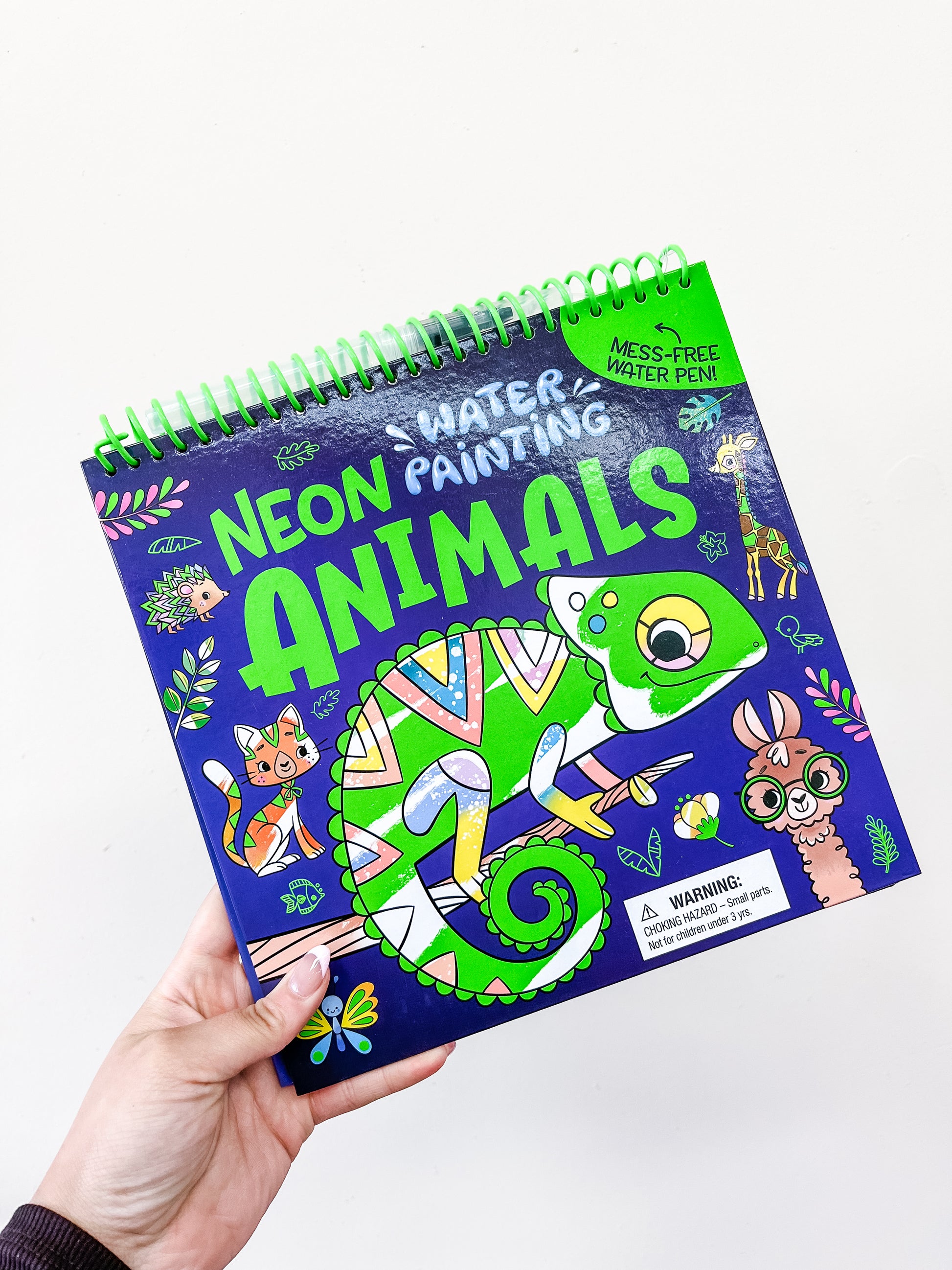 A hand holds the Water Painting: Neon Animals Book by Simon & Schuster, a spiral-bound book with vibrant animal illustrations on the cover. It offers mess-free painting fun using only water and a special pen.