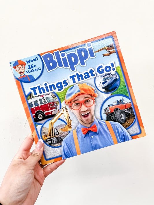 A hand holds the Simon & Schuster Blippi Things That Go! Book, featuring Blippi on the cover with images of a fire truck, excavator, airplane, boat, and monster truck. The book includes over 25 stickers.