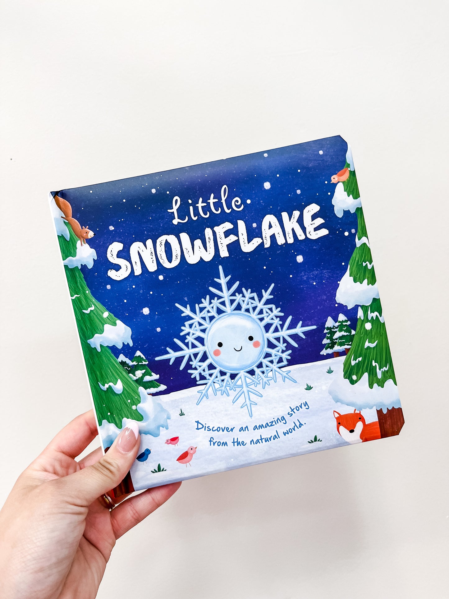 A hand holds the Little Snowflake Book by Simon & Schuster, featuring a smiling snowflake on the cover with fir trees, a fox, and a bird in a winter forest beneath a snowy night sky.