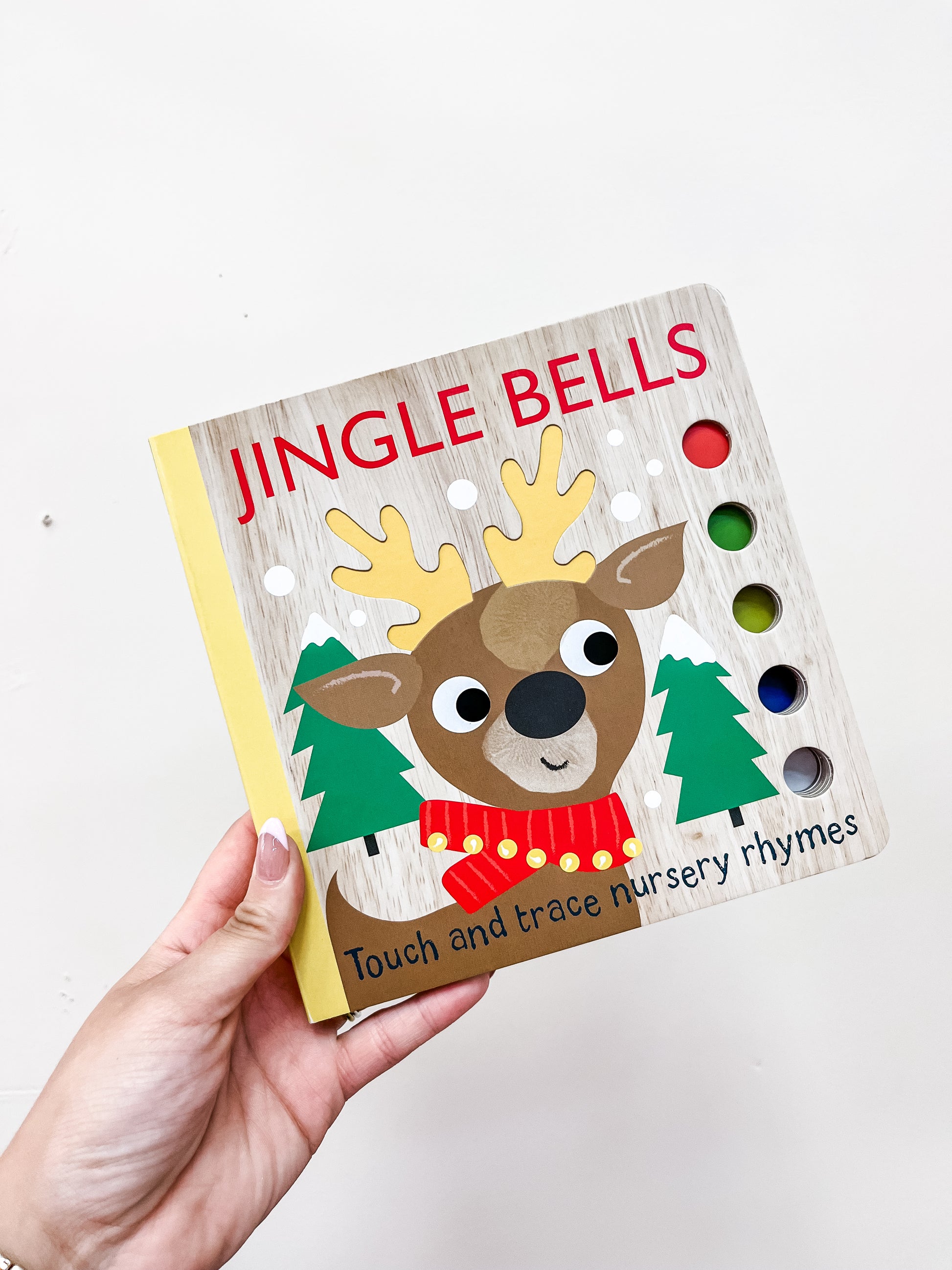 A hand holds the Simon & Schuster Jingle Bells Book, a children's board book with a cartoon reindeer, green trees, and colorful touch-and-trace holes on the cover. The subtitle reads "Touch and trace nursery rhymes.