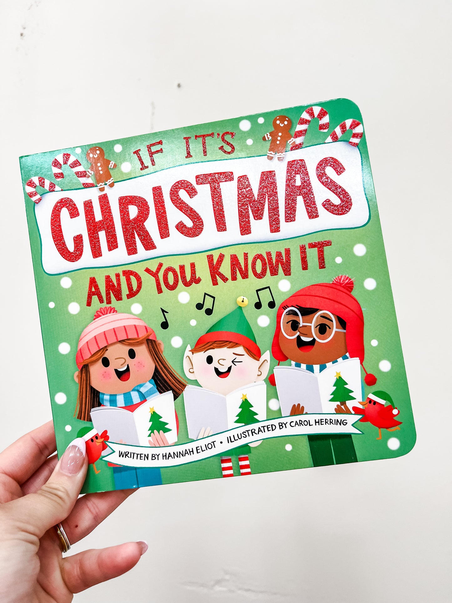 A hand holds the Simon & Schuster "If It's Christmas and You Know It Book," featuring three smiling kids in winter clothes singing with songbooks amid festive decorations on the cover.