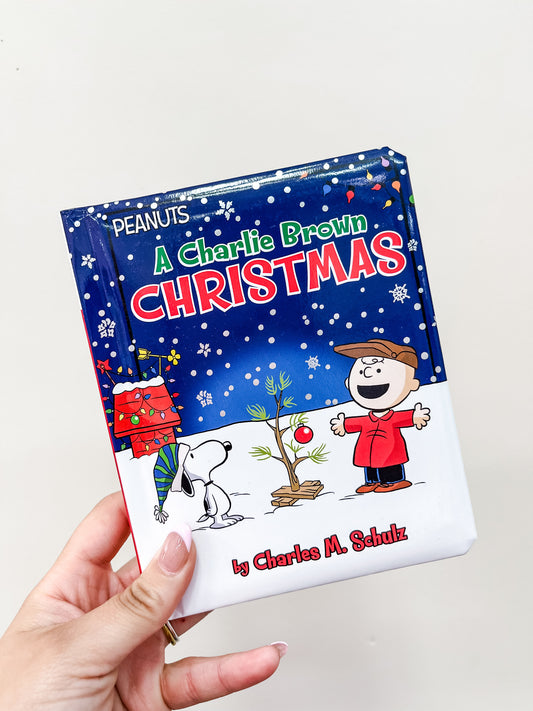 A hand holds the Charlie Brown Christmas Book by Simon & Schuster, showing Charlie Brown, Snoopy, and a small Christmas tree with snowflakes on the cover.